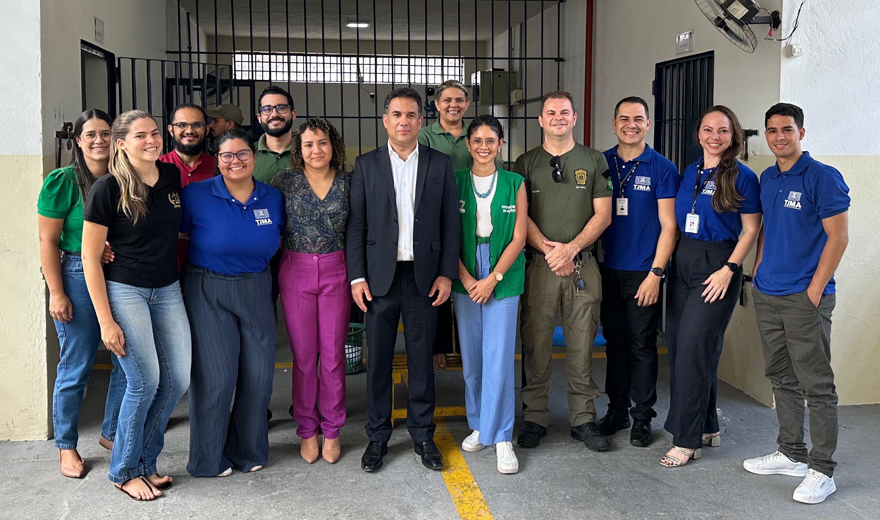 ~Foto horizontal, colorida, de grupo de vários homens e mulheres de pé, posando pra foto, em ambiente interno com paredes de cor amarela, com grades ao fundo.