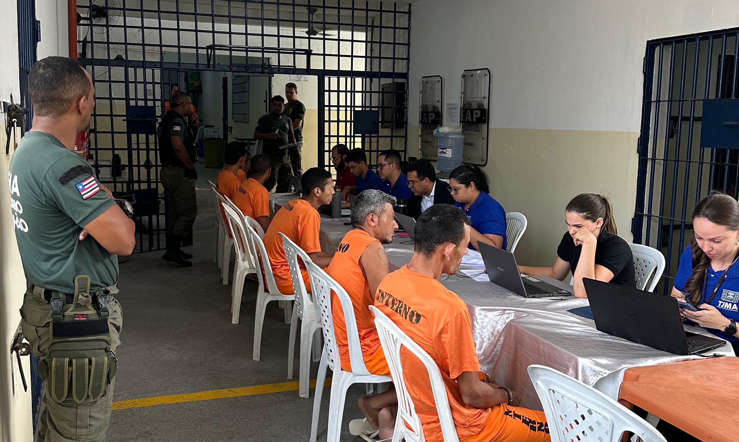 Foto horizontal, colorida, de pessoas presas, vestidas de uniforme laranja, com a palavra "INTERNO" escrita nas costas, sentadas em cadeiras brancas, diante de mesas brancas onde outras pessoas estão sentadas, diante de computadores. À esquerda, policial de pé, vestido de uniforme verde. A cena ocorre em um ambiente fechado com grades de metal ao fundo, onde há outros homens uniformizados.