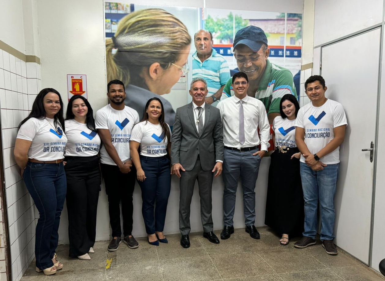 Foto horizontal, colorida, de homens e mulheres de pé, diante de parede clara com cartaz onde se vê pessoas reunidas. A maioria das pessoas usa camiseta branca, com cores azul e verde, onde se lê: Semana Nacional da Conciliação. Elas posam pra foto.