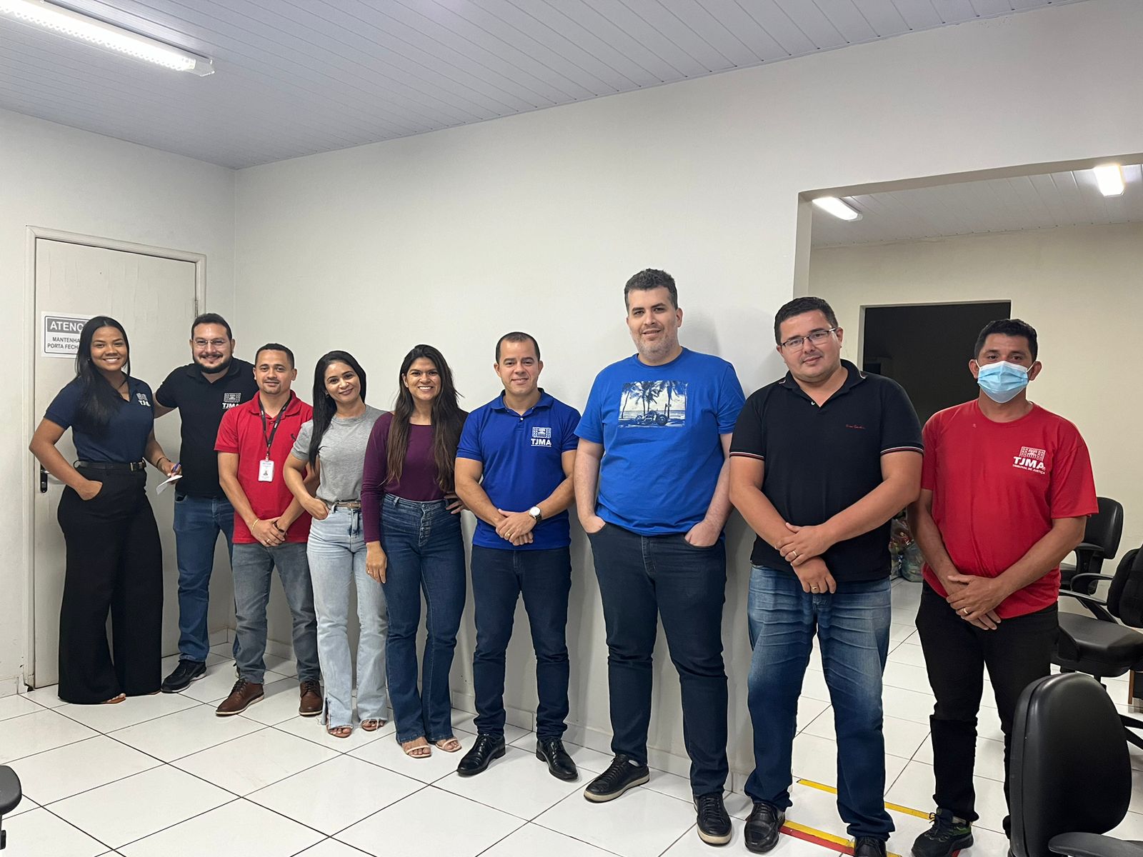 Um grupo maior de pessoas, composto por cinco homens e quatro mulheres, também está posando juntos em um ambiente de escritório. Os homens estão em trajes casuais, com alguns usando camisas polo e outros com camisetas. Duas mulheres estão de calça jeans e camisetas, e as outras duas estão vestidas de forma mais formal, uma delas usando calça social. Um dos homens na extremidade direita está usando máscara facial, o que pode indicar cuidados de saúde no ambiente.