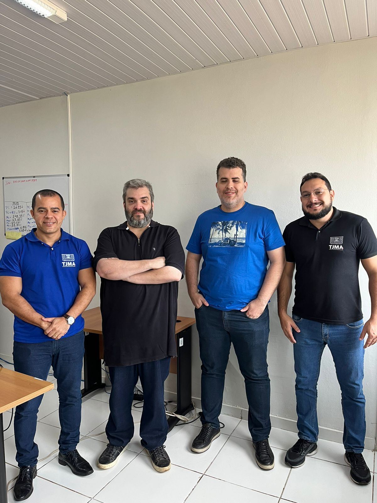 Quatro homens estão de pé próximos a mesas de escritório. Todos usam roupas casuais: dois estão com camisetas azuis, um com camiseta preta, e o outro com uma camisa polo preta. Eles parecem descontraídos e estão sorrindo.