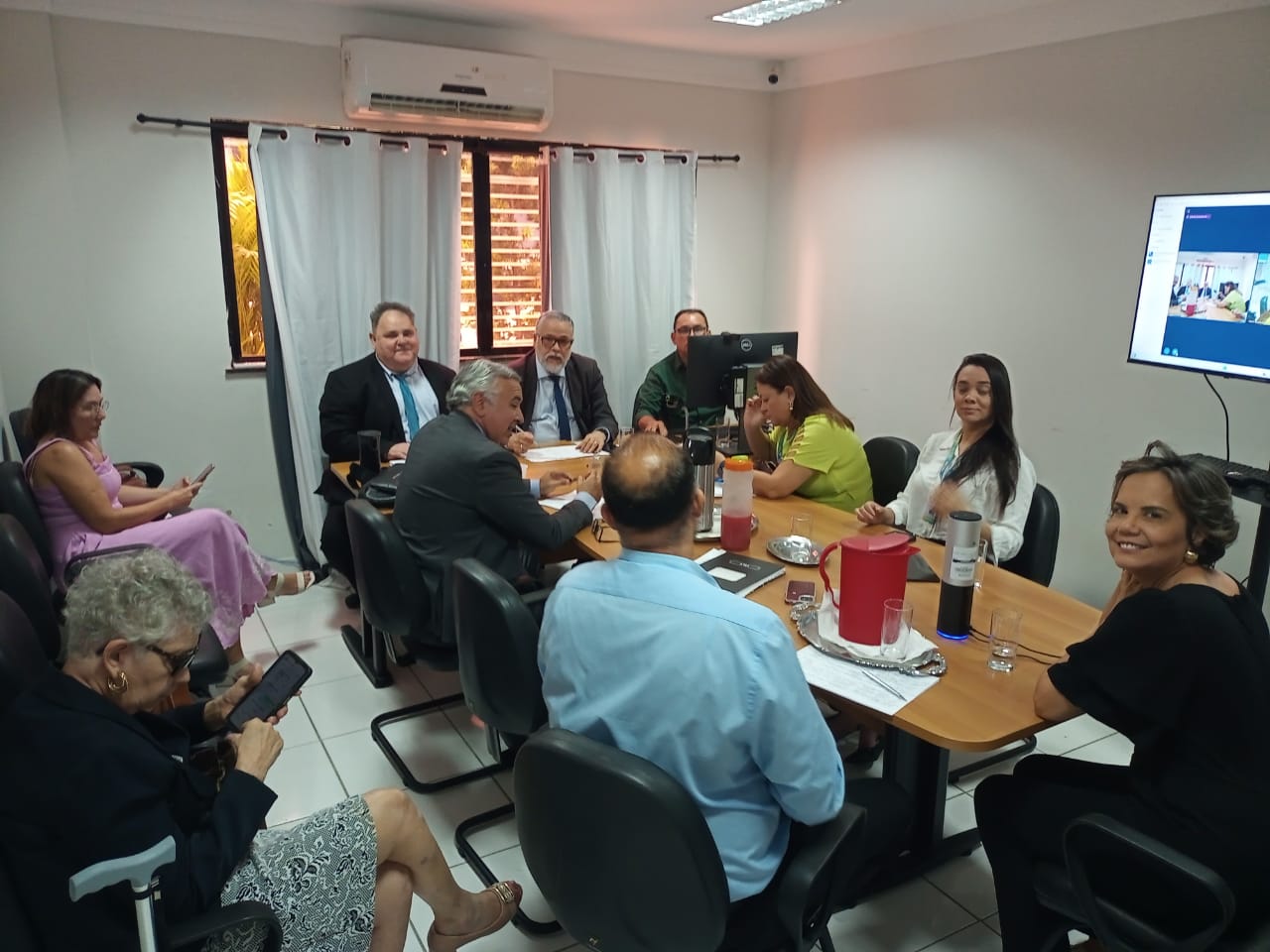 Foto horizontal, colorida, de homens e mulheres claras. Pessoas estão sentadas ao lado direito, acompanhando a reunião. Ao fundo, uma janela com cortinas e um aparelho de ar-condicionado na parede.