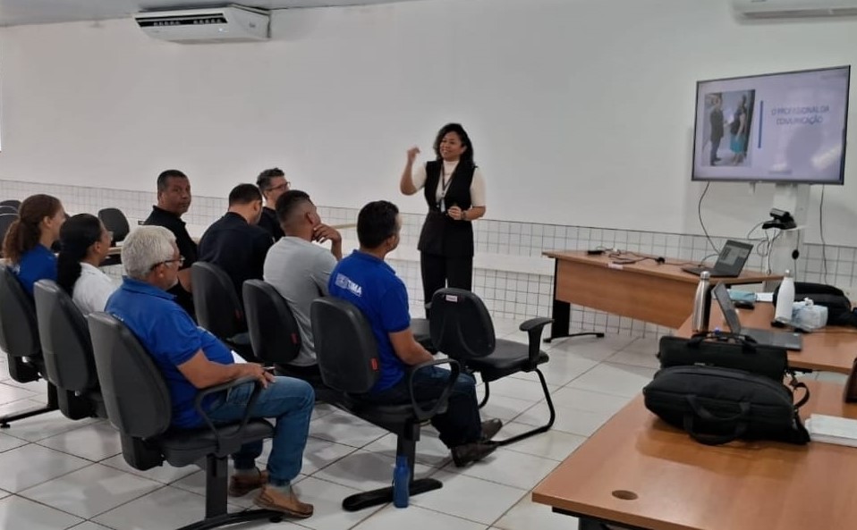 Foto horizontal, colorida, de homens e mulheres sentados em sala assistindo a uma apresentação. Há uma mulher parda, em pé na frente da sala, , gesticulando enquanto fala. Na parede à direita, há uma tela de televisão ou monitor exibindo uma apresentação com o título "O PROFISSIONAL DA COMUNICAÇÃO". Há também uma mesa com um laptop, uma garrafa de água e outros materiais de escritório. A sala é bem iluminada, com luzes no teto e um ar-condicionado montado na parede.