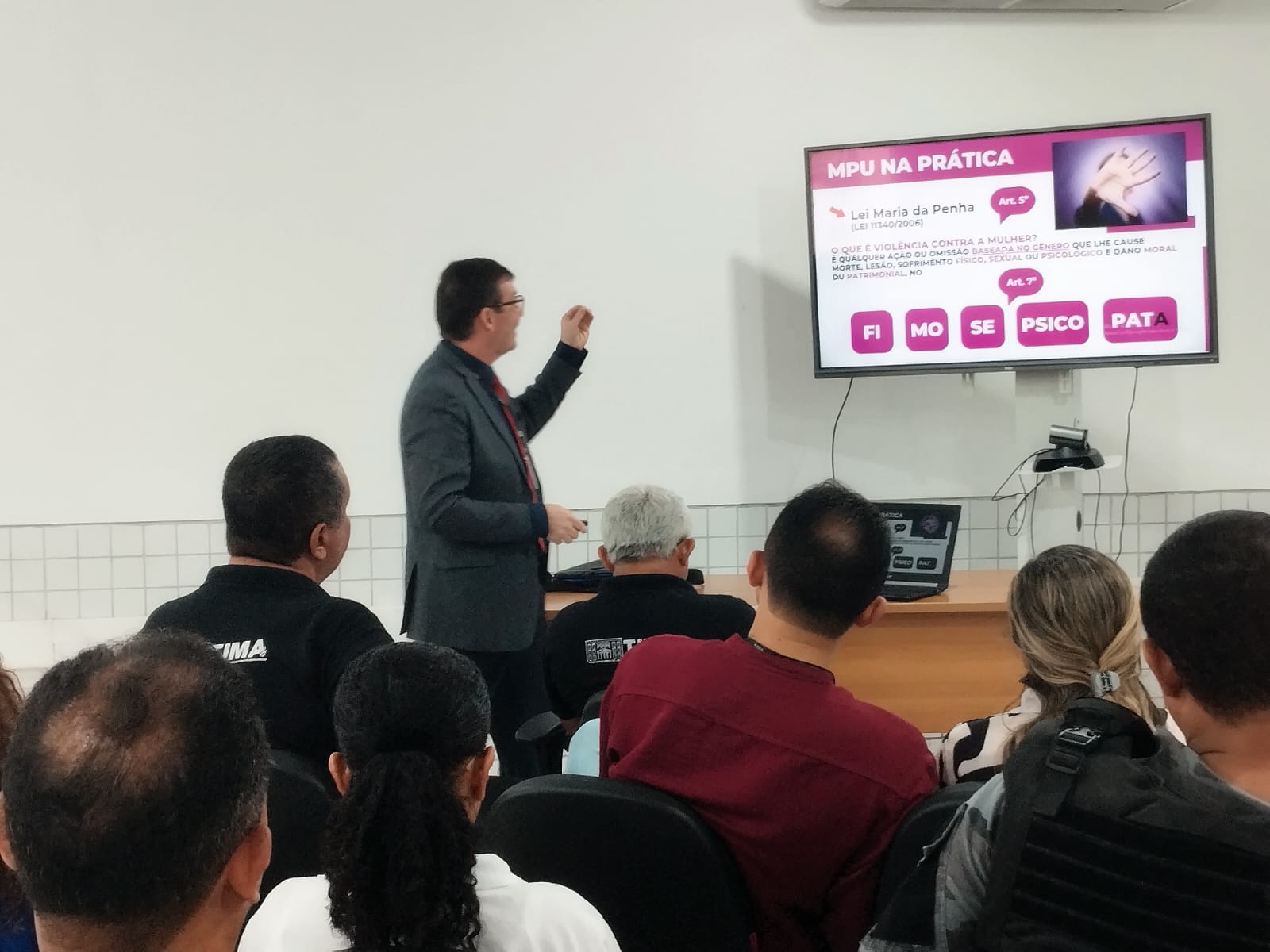 Foto horizontal, colorida, de pessoas em auditório, de costas, olhando para homem pardo, de terno escuro, que aponta para uma tela de projeção na parede, com informações sobre a "Lei Maria da Penha" (Lei 11.340/2006), com o texto:"MPU NA PRÁTICA" e menciona os artigos 5º e 7º da lei.

Abaixo do título, há uma pergunta: "O QUE É VIOLÊNCIA CONTRA A MULHER?", seguida por uma explicação sobre os tipos de violência que causam morte, lesão, sofrimento físico, sexual ou psicológico, além de danos morais ou patrimoniais. A tela também apresenta abreviações como "FI", "MO", "SE", "PSICO" e "PATA".

A plateia está sentada, prestando atenção à apresentação.

Se precisar de mais alguma coisa ou tiver outras perguntas, estou aqui para ajudar! ¿