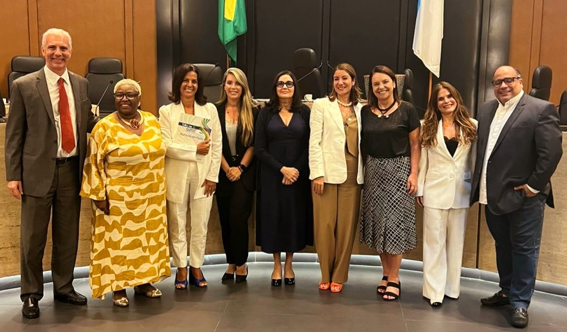 Foto horizontal, colorida, de homens e mulheres de pé, em salão de eventos., com bandeiras hasteadas ao fundo. As pessoas estão usando trajes formais e posam para foto.