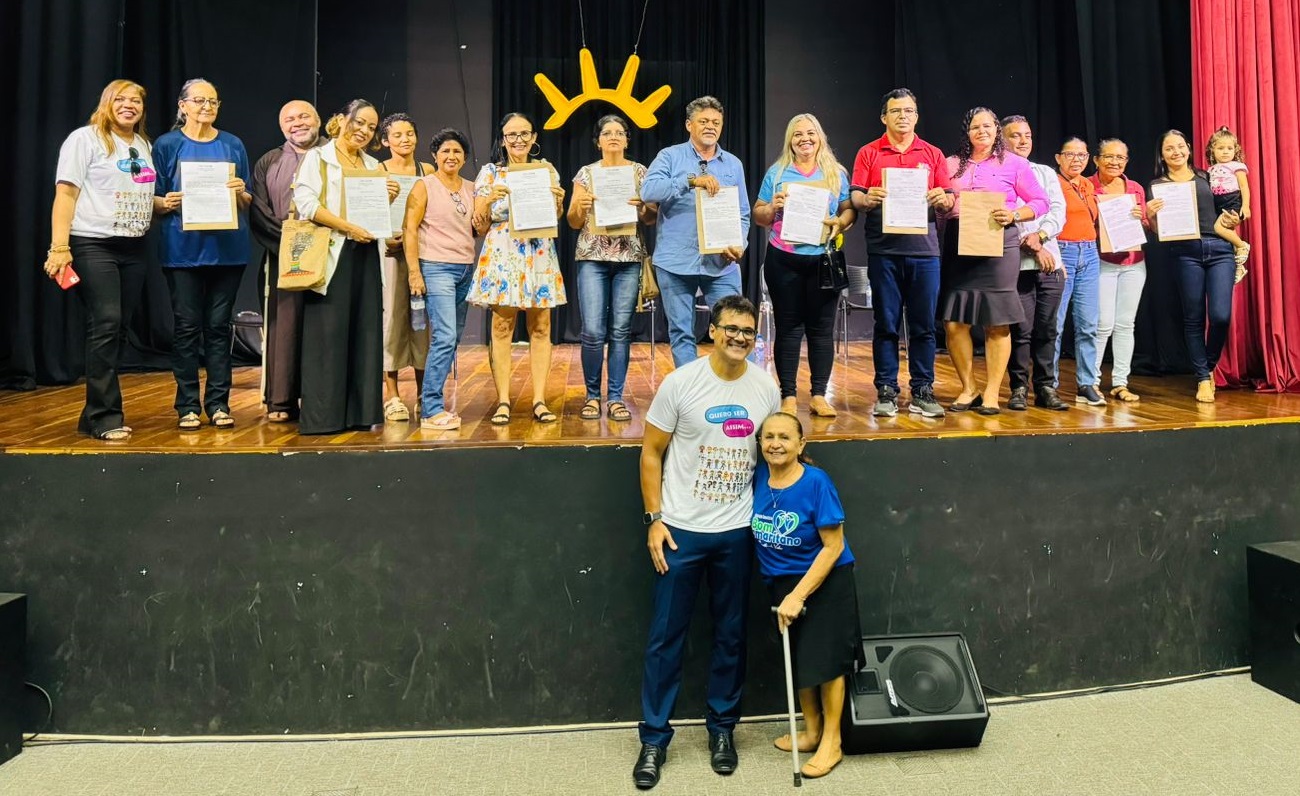 Foto horizontal, colorida, de um grupo de pessoas de pé em um palco com cortinas vermelhas. Elas seguram documentos. O palco tem um fundo preto com uma decoração amarela em forma de sol no centro. Há duas pessoas em frente ao palco, uma delas usando uma bengala.