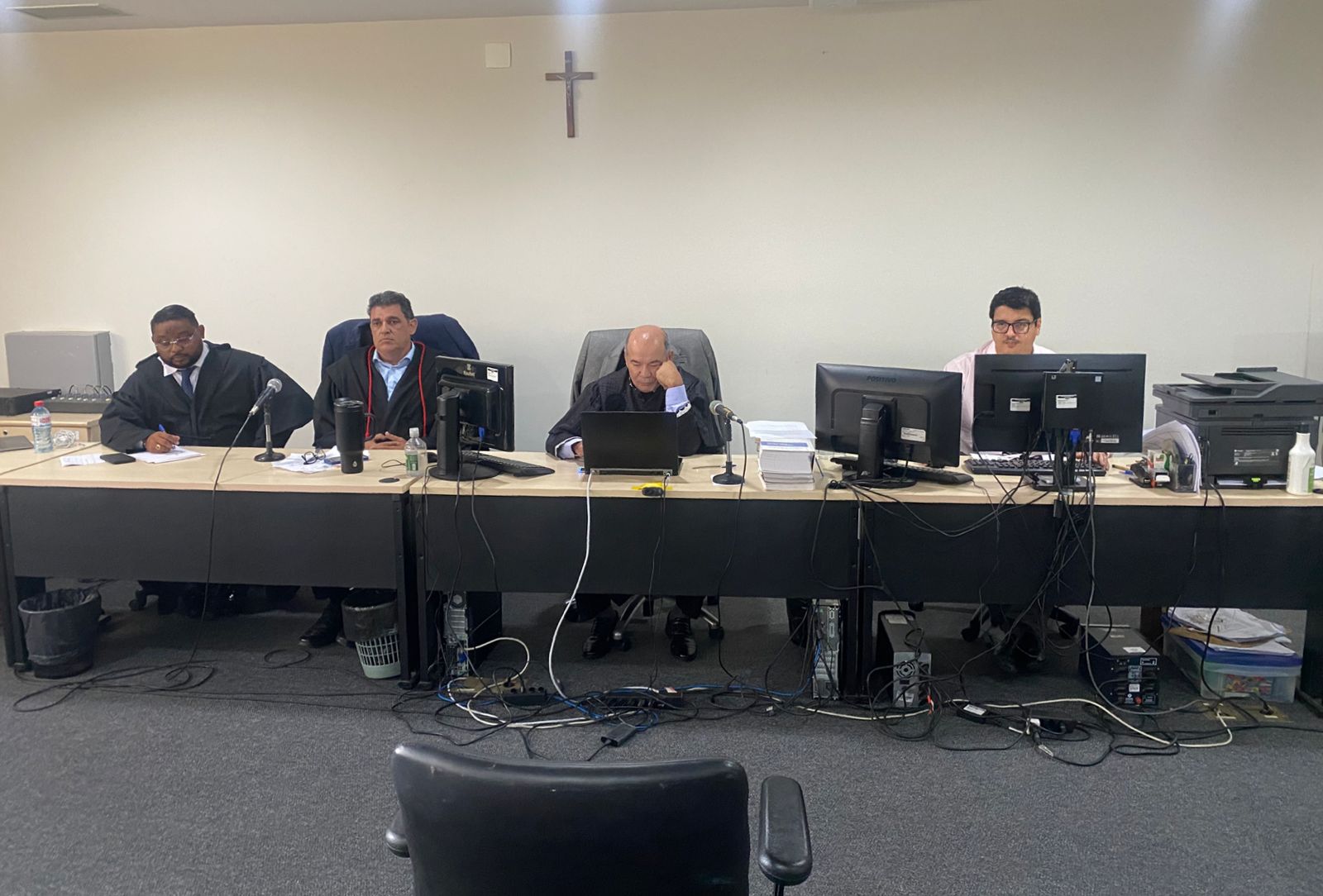 A imagem mostra uma bancada em um ambiente de audiência judicial do 1º Tribunal do Júri. Quatro pessoas estão sentadas na mesa: dois homens em togas, um deles escrevendo e outro aparenta estar em reflexão, além de dois outros profissionais utilizando computadores. No centro da parede ao fundo, há uma cruz fixada, reforçando o contexto formal e judicial. A mesa contém materiais como microfones, papéis, laptops e garrafas de água. No chão, há vários cabos conectando os equipamentos eletrônicos.