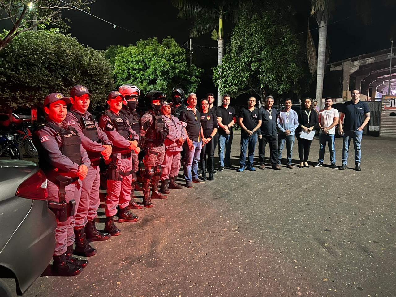 Imagem horizontal, colorida, em ambiente externo e noturno, mostra uma fileira de homens de pé, em forma de meia-lua, do lado esquerdo.Alguns deles vestem uniformes policiais.