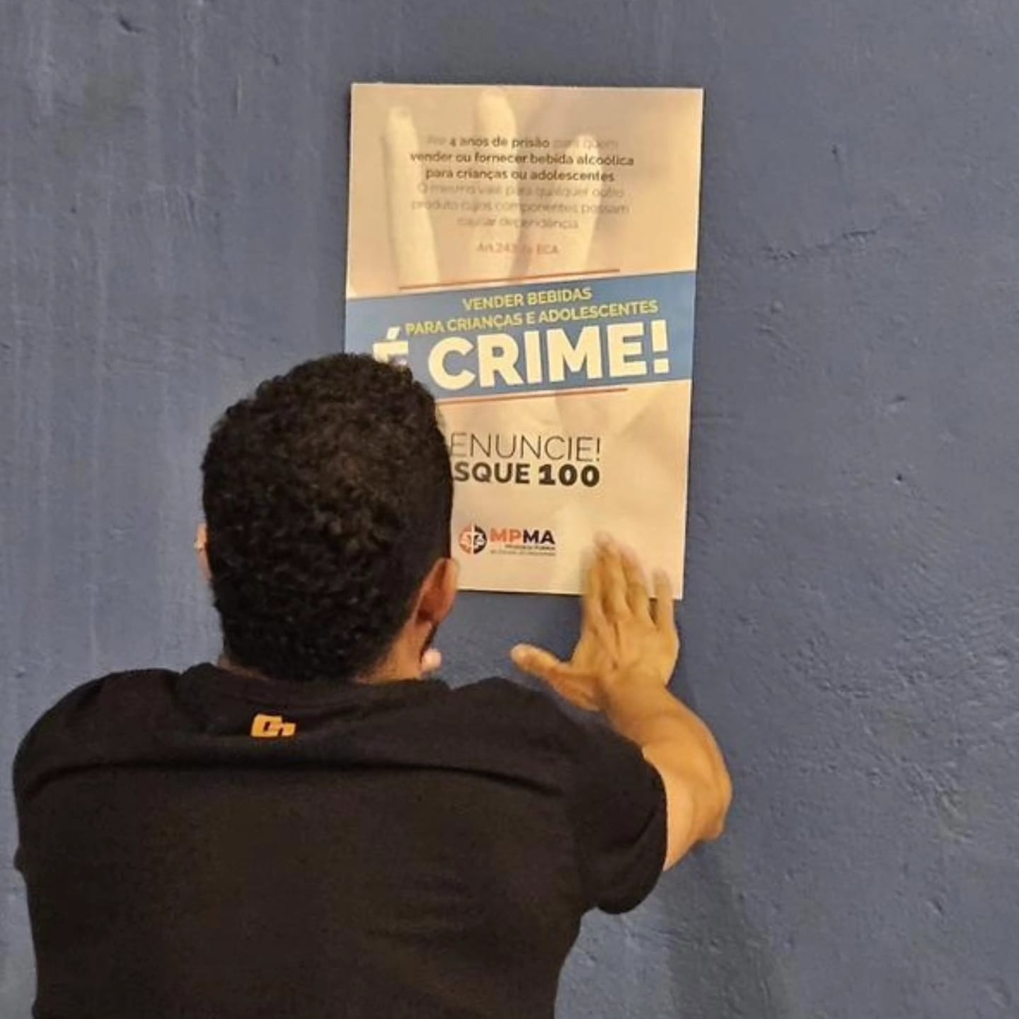 Imagem horizontal, colorida, de homem de costas, vestido de camiseta preta, fixando cartaz em parede cinza. No cartaz está escrito "É crime", "disque 100".