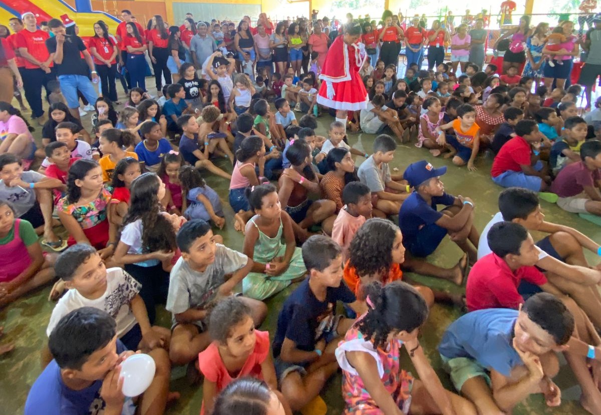 Imagem horizontal, colorida, mostra dezenas de crianças sentadas no chão em ginásio de esportes. Ao centro, uma menina vestida de "Mamãe Noel", de pé, entre as crianças.