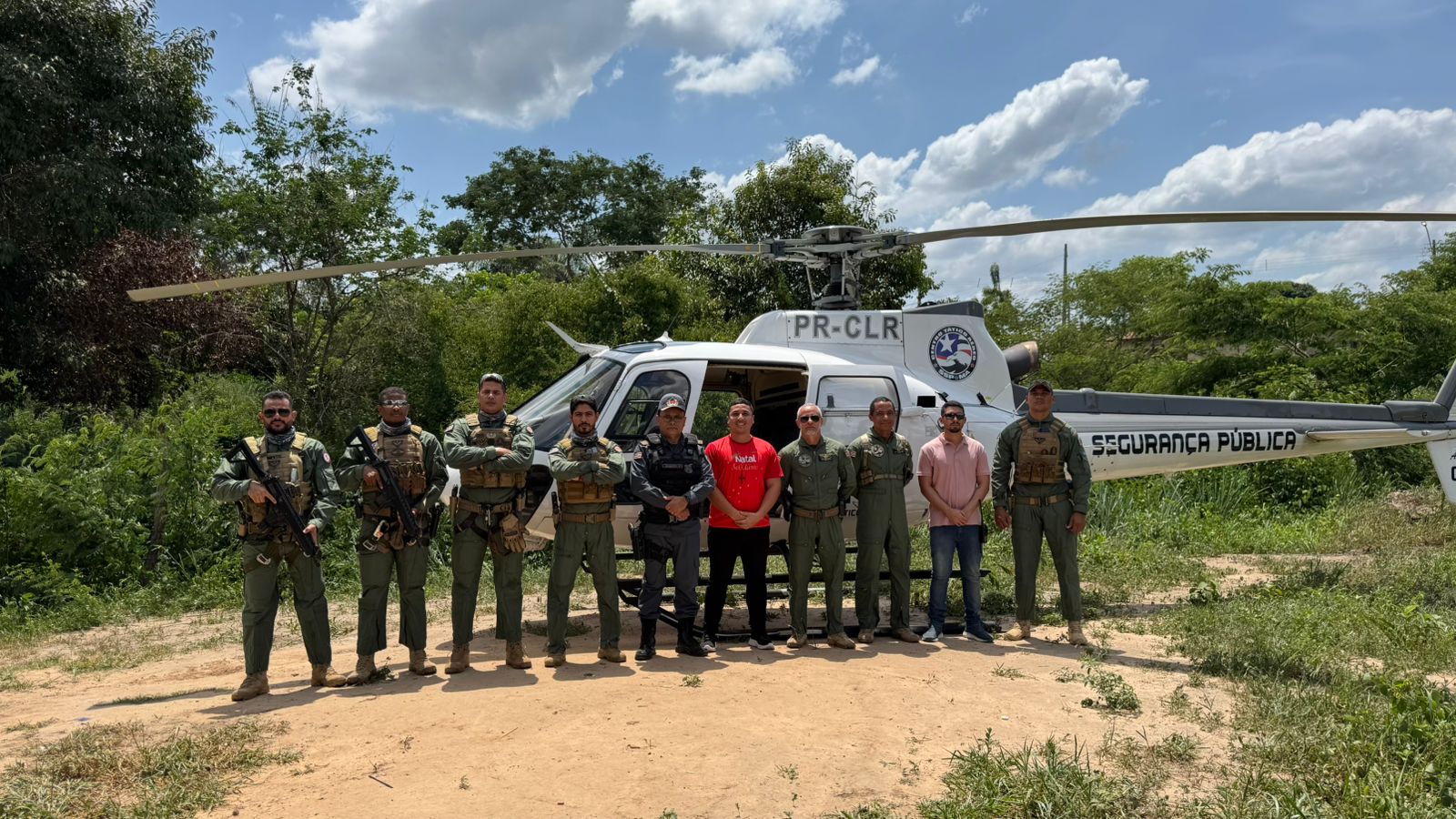 Foto horizontal, colorida, mostra 10 homens lado a lado, em área verde, diante de um helicóptero branco. Alguns deles estão vestidos com farda da polícia militar.