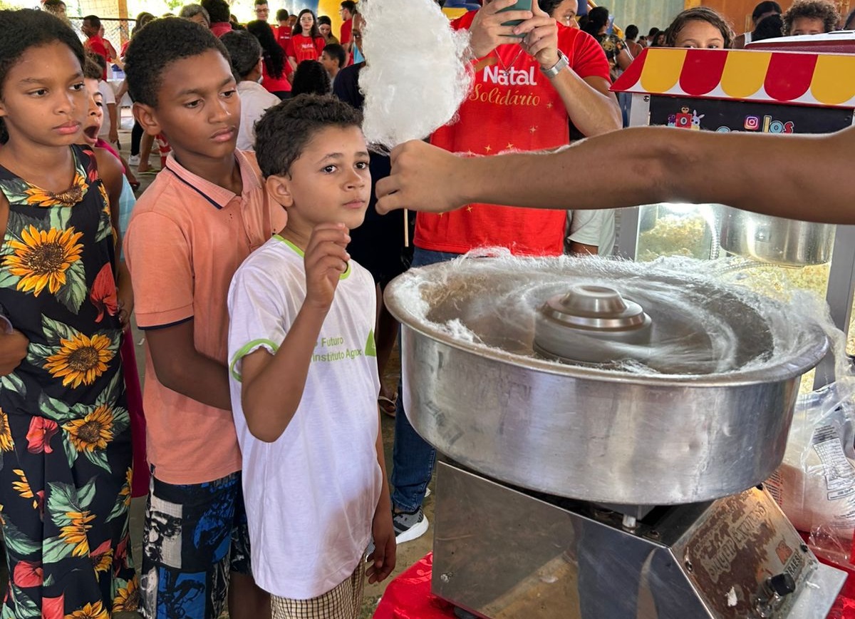 Imagem horizontal, colorida, de três crianças pardas, em fila,em ginásio de esportes, diante de panela de alumínio, recebendo algodão-doce.