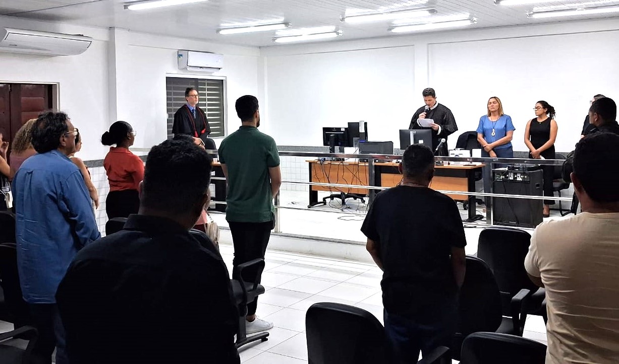 Imagem horizontal, colorida,  mostra uma sala de tribunal com várias pessoas em pé. Na frente da sala, há uma mesa com computadores e equipamentos eletrônicos. Atrás da mesa, há quatro pessoas em pé, uma delas segurando um papel e aparentemente falando. As outras pessoas na sala estão de pé, voltadas para a frente. A sala é bem iluminada e possui ar-condicionado.