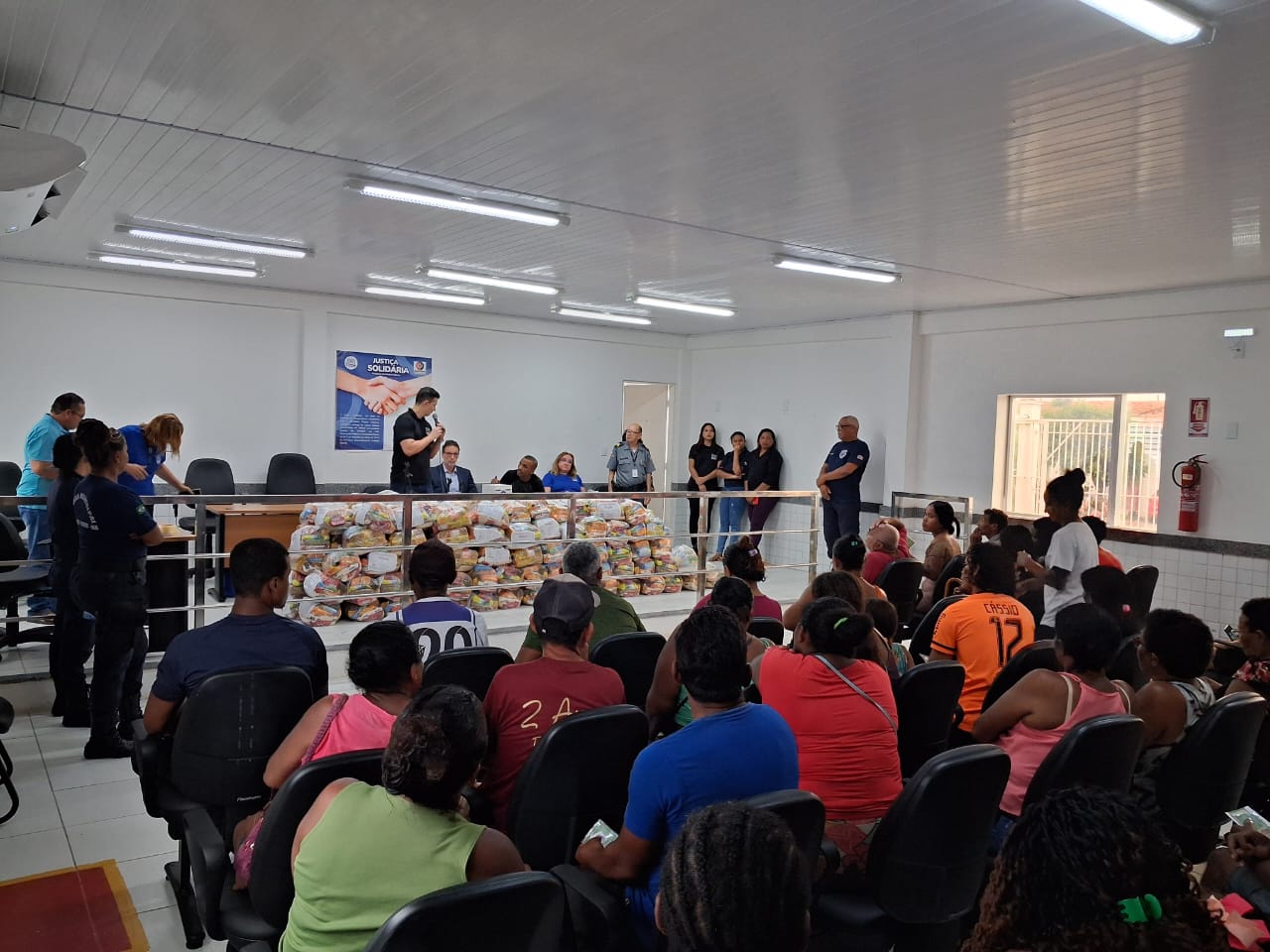 Imagem horizontal, mostra um evento comunitário em salão bem iluminado. Há homens e mulheres sentadas em fileiras de cadeiras voltadas para um grupo de indivíduos em pé e sentados na frente da sala. Dois homens em bancada lideram o evento. Há pilhas de pacotes, com alimentos para doações em mesas na frente. Um homem está falando ao microfone, dirigindo-se ao público. Um cartaz na parede que diz "Ação Solidária" com a imagem de duas mãos se apertando. A sala tem paredes brancas, piso de azulejo e iluminação fluorescente. Há também um extintor de incêndio montado na parede à direita.

Se precisar de algo mais ou tiver outra imagem, me avise! ¿