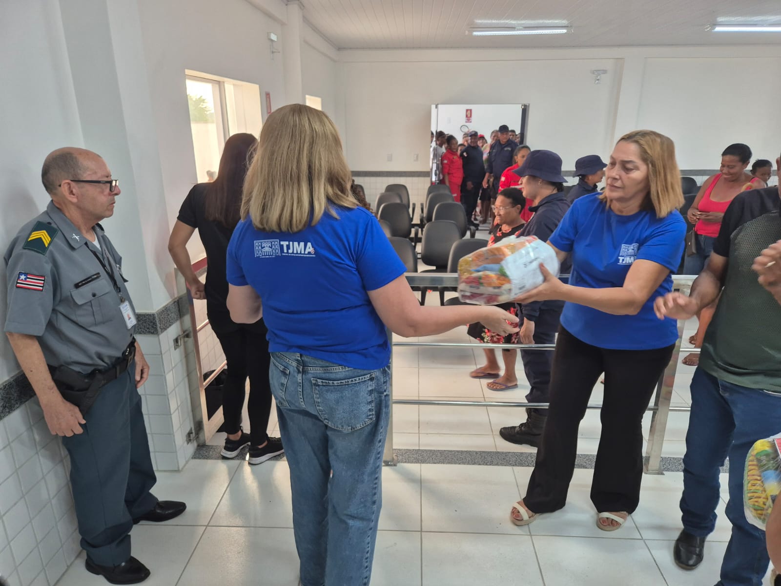 IMAGEM HORIZONTAL, COLORIDA, MOSTRA DUAS MULHERES BRANCAS, DE CABELOS LOUROS, VESTIDAS DE CAMISETA AZUL ANIL, DE PÉ, ENTRE OUTRAS PESSOAS EM AUDITÓRIO DE PAREDES BRANCAS. UMA DELAS, DE FRENTE, ENTREGA UMA CESTA BÁSICA DE ALIMENTOS PARA OUTRA. À ESQUERDA, UM, HOMEM DE PÉ, VESTIDO DE UNIFORME POLICIAL, OBSERVA.
