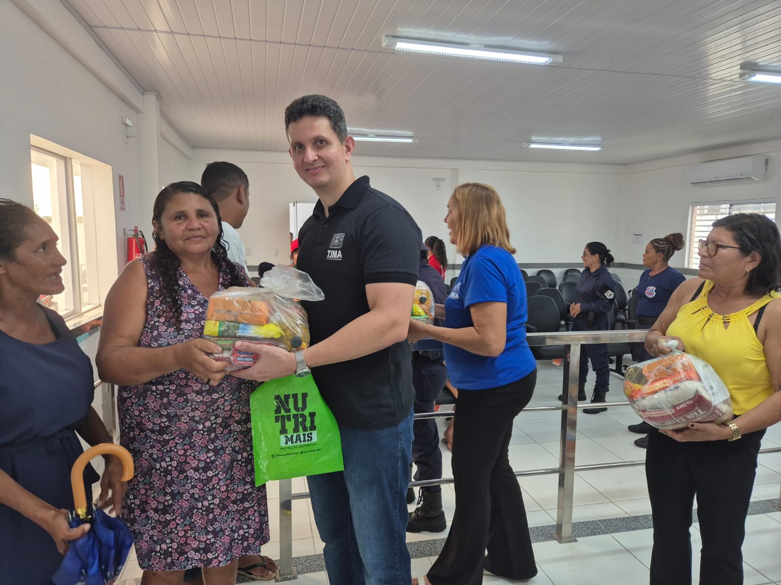 Imagem horizontal, colorida, mostra um grupo de pessoas em sala com paredes e piso brancos. Várias pessoas estão em fila, e algumas estão segurando sacos de alimentos ou suprimentos. Uma pessoa com uma camisa preta entrega um saco de alimentos a outra pessoa. Os sacos contêm vários itens alimentícios, e um dos sacos tem o texto "NUTRI MAIS" impresso nele. Também há pessoas ao fundo, algumas vestindo uniformes azuis.