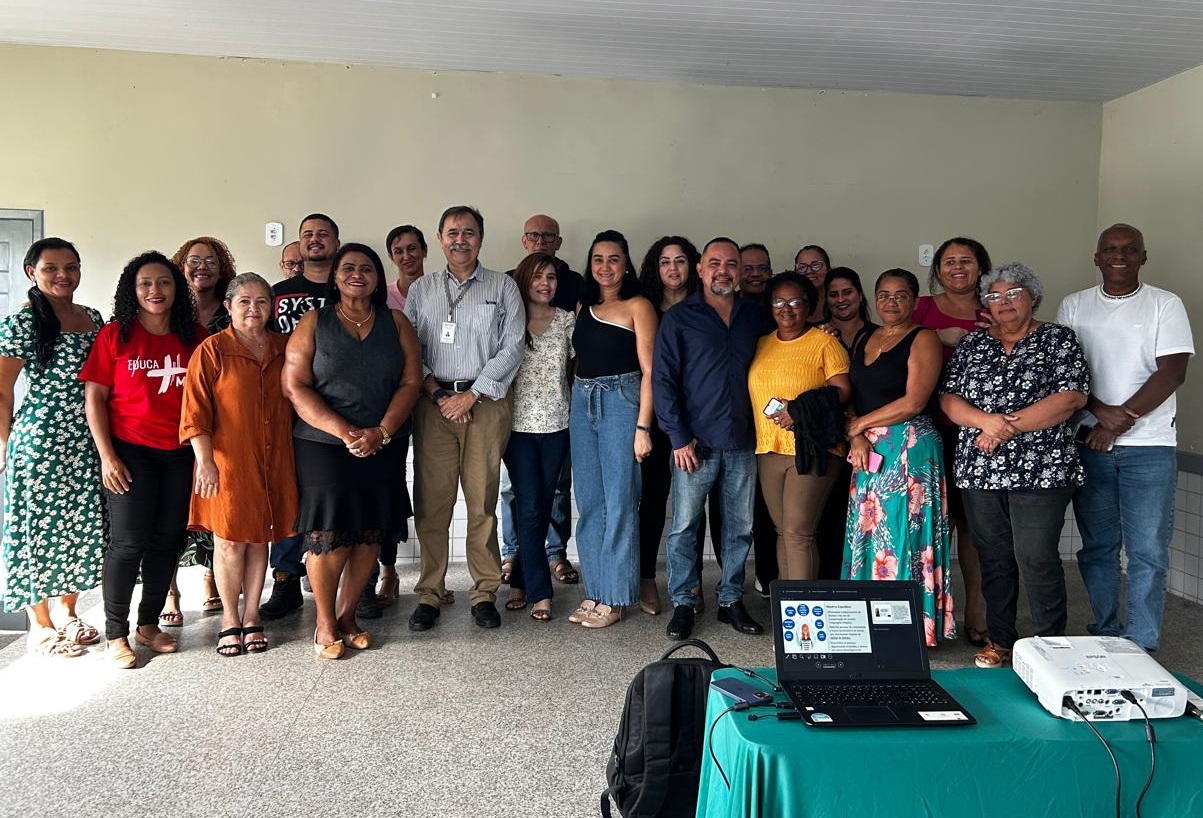 Foto horizontal, colorida, de homens e mulheres em roupas formais, de pé, junto a parede clara. Em primeiro plano, à direita, mesa com computador projetando tela.