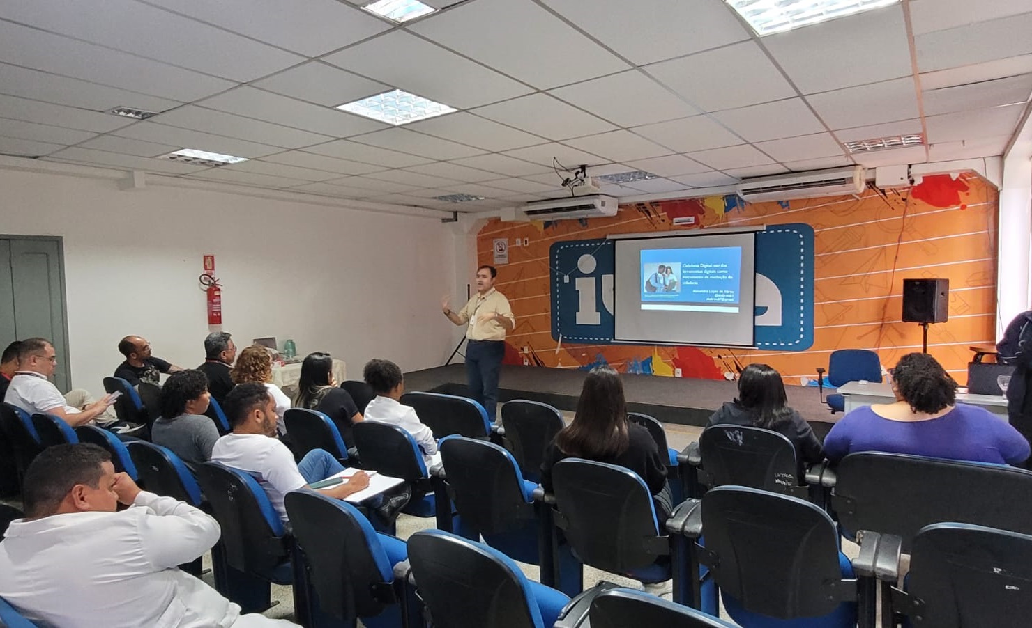 Foto horizontal, colorida, mostra um grupo de pessoas sentadas em uma sala de aula ou sala de conferências, voltadas para um apresentador que está de pé próximo a uma tela de projeção. O apresentador está gesticulando com uma das mãos enquanto fala. A tela de projeção exibe um slide com texto e uma imagem de duas pessoas. O slide lê: "Cidadania Digital: uso das tecnologias digitais e a construção da cidadania no contexto da sociedade da informação e do conhecimento." Abaixo disso, diz: "Apresentação: Ludmila de Paula Coordenadora de Projetos de Inclusão Digital." A sala possui cadeiras azuis dispostas em fileiras, e as paredes são decoradas com um mural colorido. Há um extintor de incêndio montado na parede à esquerda.