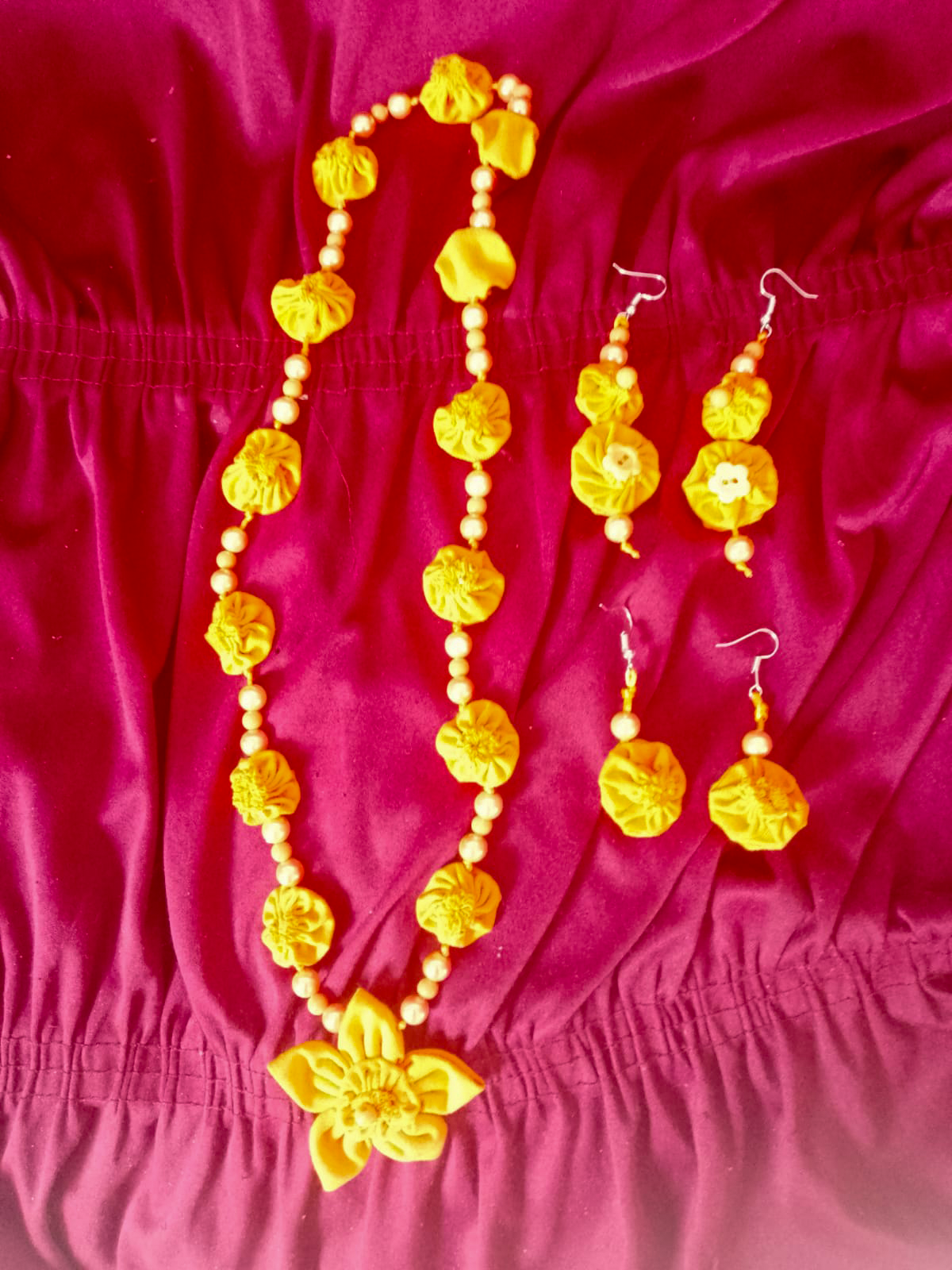 Conjunto de acessórios composto por um colar e dois pares de brincos feitos com tecido amarelo e miçangas. O colar tem flores de tecido intercaladas com pequenas contas arredondadas e um pingente central em forma de flor. Os brincos seguem o mesmo estilo, com flores de tecido e detalhes de contas. As peças estão dispostas sobre um tecido roxo franzido.