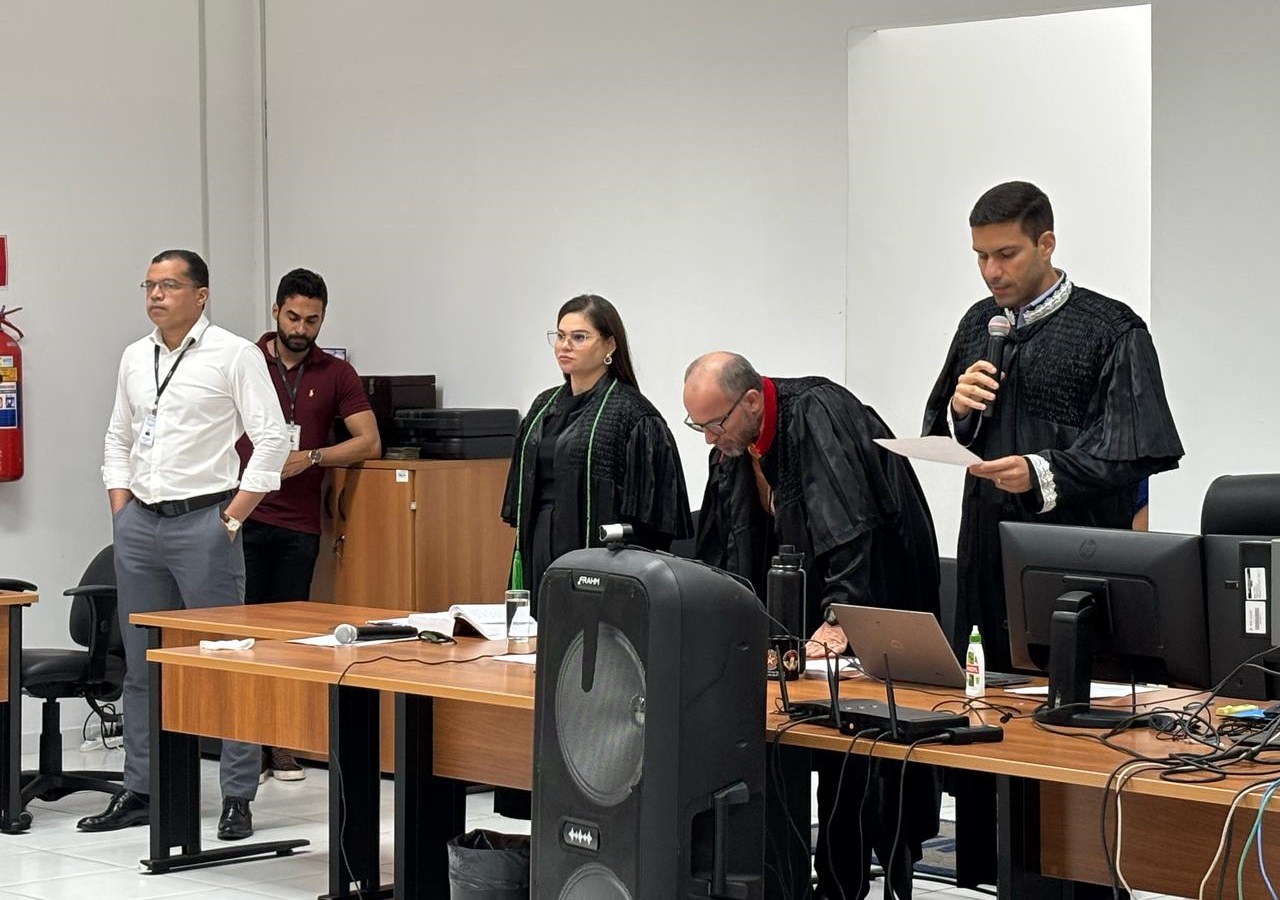 Foto horizontal colorida, mostra um grupo de pessoas reunidas em uma sala de Tribunal do Júri. Existem cinco indivíduos visíveis, sendo que três deles estão vestidos com togas pretas, sugerindo que possam ser juízes ou funcionários. Um dos indivíduos com toga está falando ao microfone e segurando um papel, possivelmente lendo ou fazendo um anúncio. Os outros dois indivíduos de toga estão perto de uma mesa com vários itens sobre ela, incluindo um microfone, um laptop e alguns documentos. Dois outros indivíduos estão de pé à esquerda, um vestindo uma camisa branca e o outro uma camisa marrom, ambos observando a cena. A sala tem paredes brancas, um extintor de incêndio montado na parede e mesas de madeira com equipamentos de escritório.
