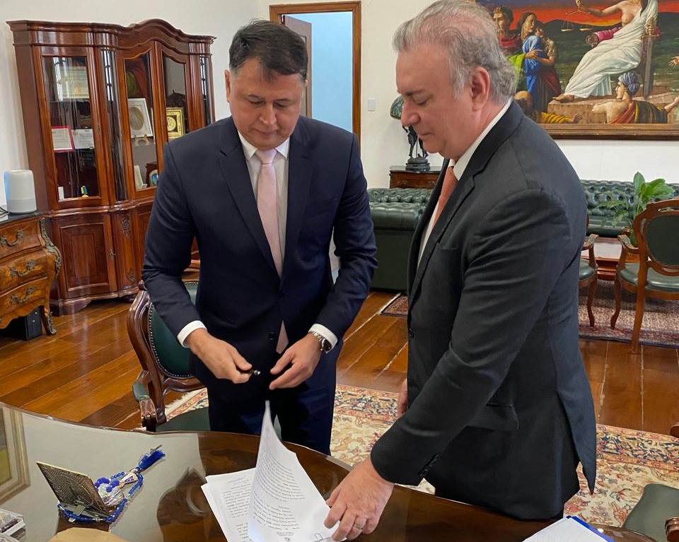 Foto horizontal, colorida, de dois homens de pé, vestidos de terno azul escuro. O homem da direta, branco, de cabelos grisalhos, está de lado e segura um documento sobre uma mesa. O homem da esquerda, branco, cabelos pretos, segura uma caneta e olha para o documento. Ao fundo, estante de madeira escura com livros, sofá verde tela colorida na parede branca.