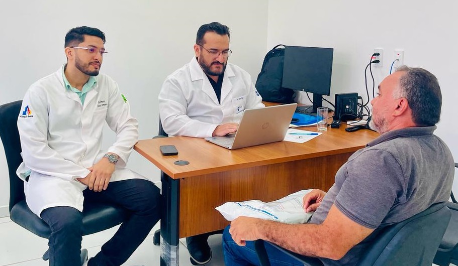 Foto horizontal, colorida, mostra três homens em um ambiente que parece ser um consultório ou escritório médico. Dois deles estão vestidos com jalecos brancos, sugerindo que sejam médicos ou profissionais de saúde. Eles estão sentados à mesa, onde há um laptop aberto. O terceiro homem, que parece ser um paciente, está sentado de frente para eles.

A sala tem paredes brancas e uma luminária no teto. Há uma mesa de madeira com um laptop, alguns papéis e outros objetos. Os dois homens de jaleco estão sentados lado a lado, enquanto o paciente está sentado em uma cadeira do outro lado da mesa.