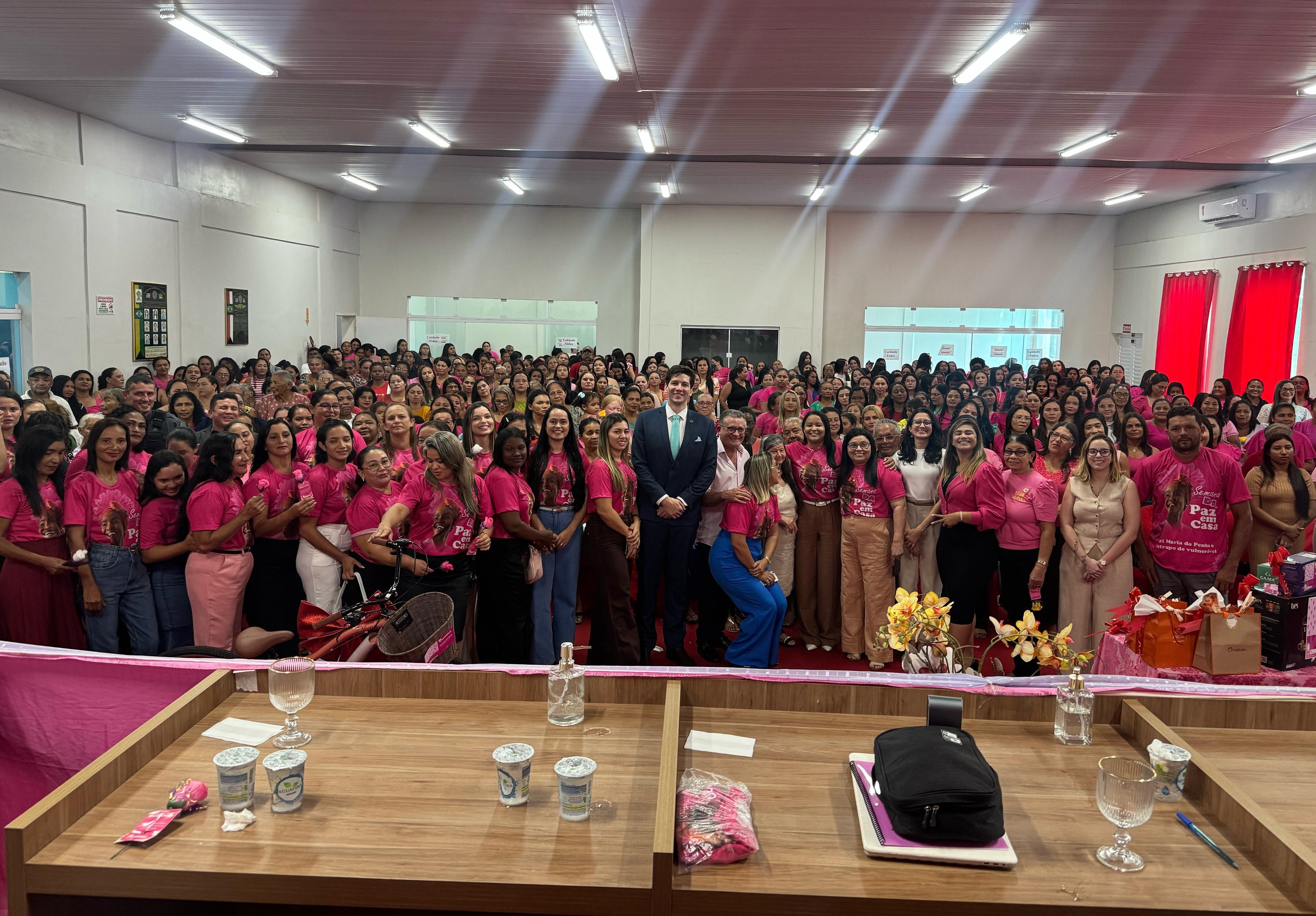 Foto horizontal, colorida, mostrando um grande grupo de pessoas reunidas em um salão espaçoso. A maioria das pessoas veste camisetas rosa, sugerindo um evento com tema de conscientização ou apoio a uma causa específica. No centro do grupo, há um homem vestindo terno azul, ladeado por outras pessoas. A composição da foto é centralizada, com o grupo de pessoas ocupando a maior parte do quadro. Em primeiro plano, há duas mesas de madeira com objetos como copos, garrafas e um notebook preto. O salão possui paredes brancas e um teto com iluminação embutida. Ao fundo, cortinas vermelhas e uma porta com uma placa.