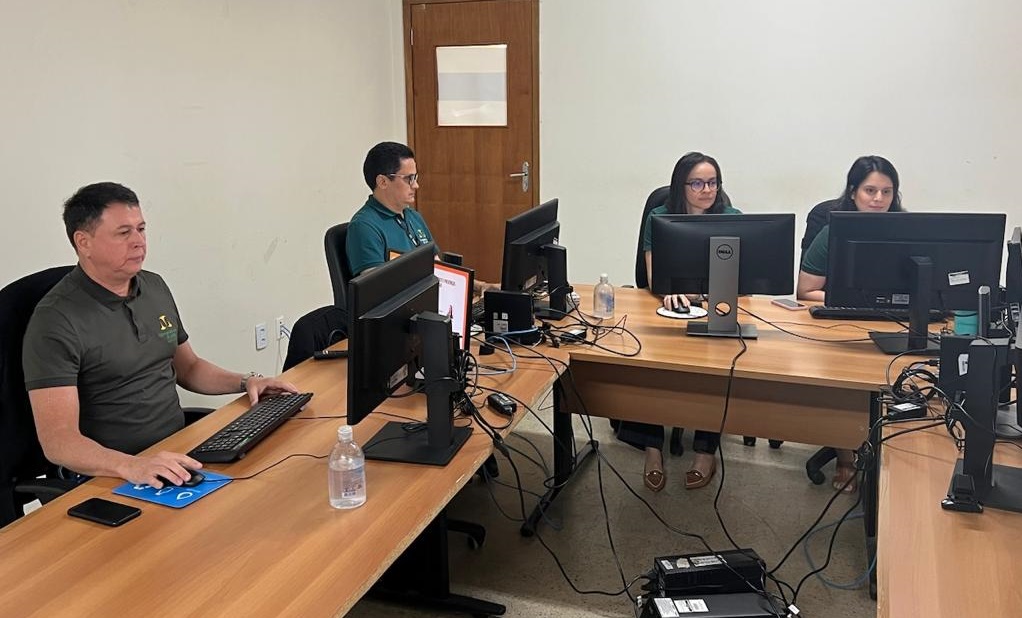 Foto horizontal, colorida, de dois homens e duas mulheres sentados diante de mesas de madeira, em formato de "U", diante de computadores. Ao fundo, parede branca, com uma porta de amdeira.