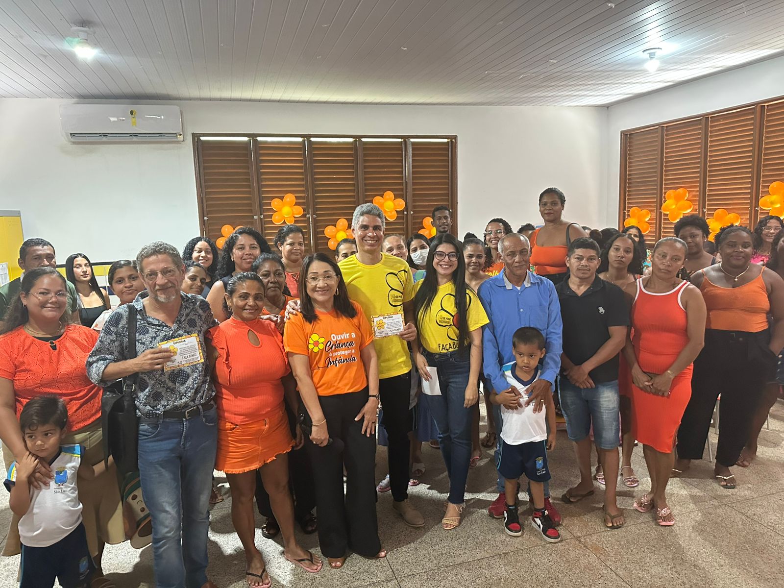 A imagem mostra um grande grupo de pessoas, incluindo homens, mulheres e crianças.

Algumas pessoas estão com camisetas amarelas e laranjas com mensagens sociais (como "Ouvir a criança é proteger a infância").

Há balões laranja na decoração.

O ambiente é informal, com muitas expressões sorridentes, sugerindo uma ação comunitária, roda de conversa ou evento de conscientização.