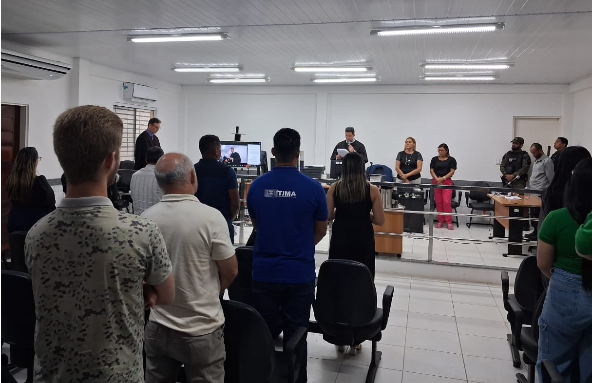 Foto horizontal, colorida, mostra o interior de uma sala, provavelmente um tribunal ou auditório, onde várias pessoas estão de pé e atentas.

No centro da imagem, um grupo de pessoas está virado de costas para o observador, olhando para a frente da sala. À frente, há uma bancada com algumas pessoas sentadas ou em pé, e uma delas, vestida de preto, parece estar lendo ou falando em um microfone. Há também um monitor visível no centro da frente da sala.

A sala tem teto branco com iluminação fluorescente e paredes brancas. Há cadeiras escuras dispostas em fileiras, e a maioria das pessoas presentes parece ser adulta.