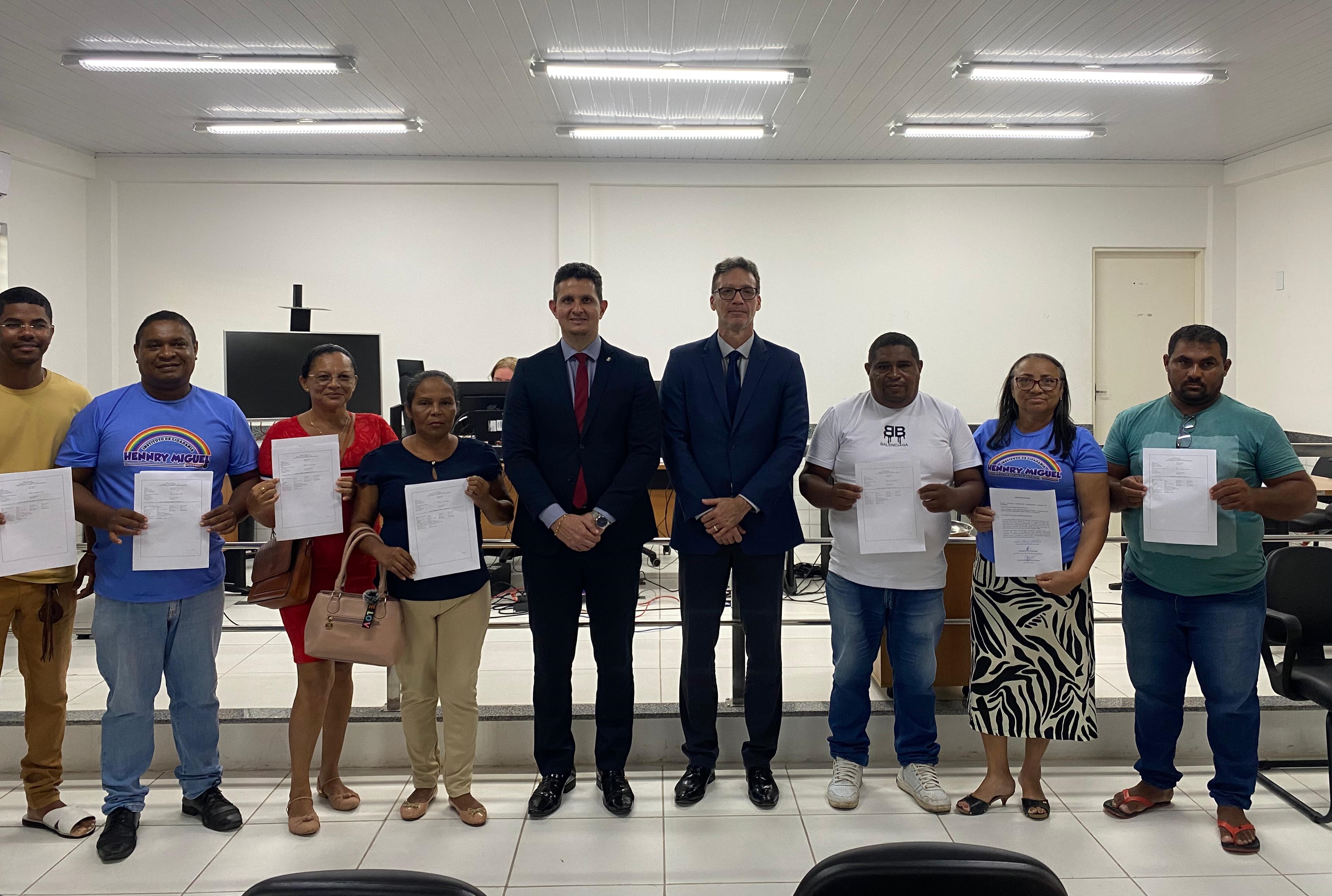 Foto horizontal, colorida, mostra um grupo de nove pessoas, entre homens e mulheres, em cerimônia, de pé, posando para uma foto. Há sete indivíduos em primeiro plano, exibindo papéis que parecem ser documentos ou certificados, e dois homens de terno escuro ao centro.

O grupo está em um auditório, com paredes brancas, iluminação fluorescente no teto e algumas cadeiras escuras visíveis na parte inferior da imagem. No fundo, à esquerda, é possível notar um monitor.