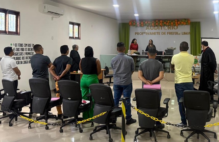 Homem condenado por homicídio em Bequimão está foragido Pergunta respondida com sucesso
A imagem mostra um auditório durante o que parece ser uma cerimônia ou reunião. O foco principal está em um grupo de sete homens e uma mulher, vistos de costas, em pé diante de várias cadeiras de escritório vazias. A maioria das cadeiras está voltada para a frente, e algumas estão encadeadas com uma fita amarela, possivelmente para delimitar o espaço ou indicar que não devem ser usadas.
Ao fundo, há um palco elevado com uma mesa comprida. Três pessoas estão sentadas atrás da mesa: duas mulheres no centro e um homem vestido com o que parece ser uma beca preta (típica de juízes, advogados ou formandos) à direita. Há também um homem de beca preta em pé no canto direito do palco, olhando para a plateia.
No centro do palco, uma placa branca com letras verdes e laranjas diz "AUDITÓRIO PROFª IVANETE DE FREITAS LIMA".
