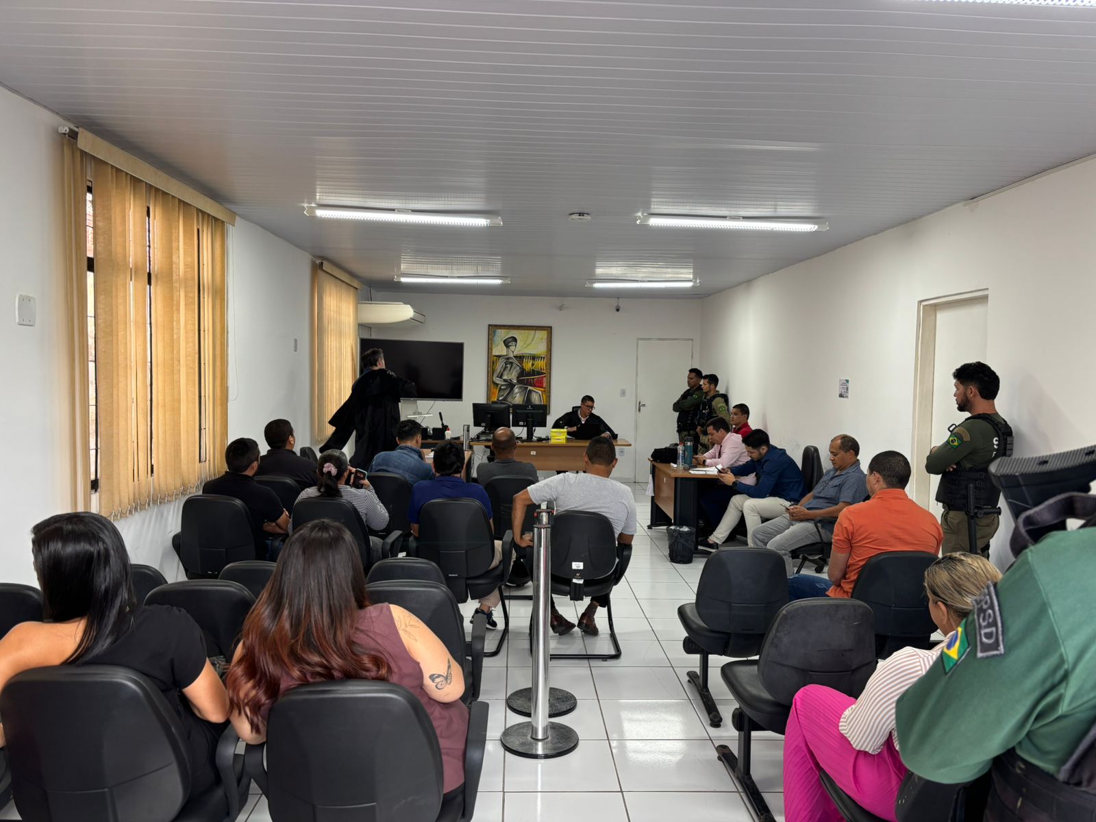 A imagem mostra uma sala de audiência judicial. O ambiente é simples, com paredes brancas e piso de cerâmica branca. Várias pessoas estão sentadas, tanto do lado do público quanto em frente à mesa do juiz, indicando que o julgamento está em andamento.

Ao fundo, há um juiz sentado à mesa, acompanhado por outras pessoas, possivelmente servidores ou membros do Ministério Público e da defesa. Um homem de toga está de pé, provavelmente um advogado ou promotor, participando da audiência. Atrás deles, vê-se uma pintura colorida e uma TV de tela plana.

À direita da imagem, há agentes de segurança uniformizados e armados, reforçando a seriedade do processo. Do lado esquerdo, pessoas do público acompanham o julgamento, algumas com roupas casuais. O ambiente está bem iluminado com luzes fluorescentes e há grandes janelas com persianas fechadas do lado esquerdo da sala.

A disposição da sala e a presença das forças de segurança indicam que se trata de um julgamento de natureza criminal.