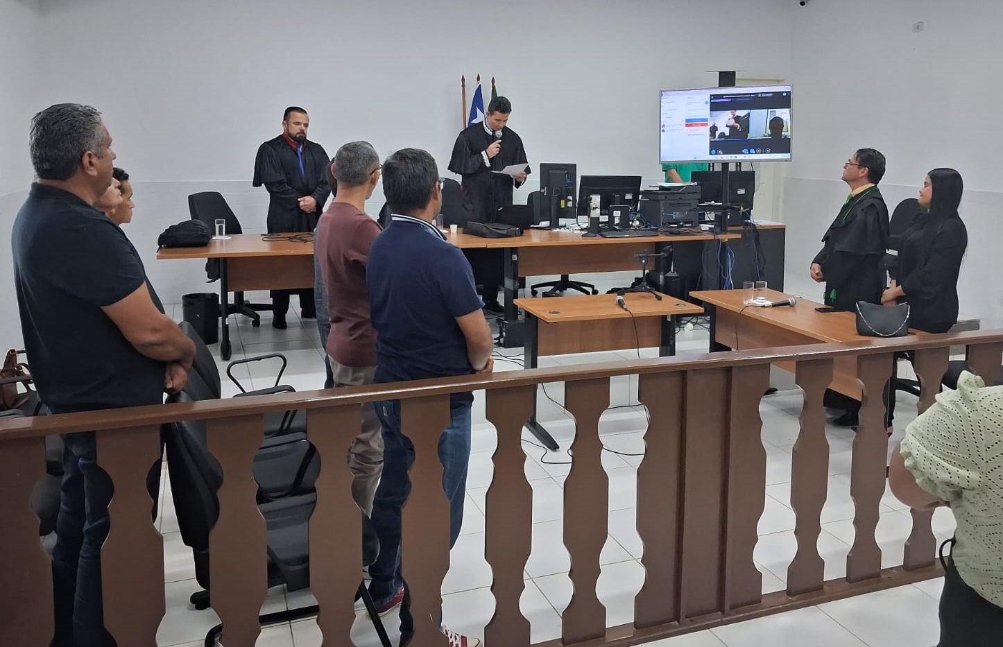 Foto horizontal, colorida, mostra sessão de Tribunal do Júri, em sala com paredes, teto e chão branco. Do lado direito da bancada, tem um monitor de TV pregado na parede com projeção em tela.

Atrás da bancada, há várias pessoas de pé, em trajes formais, escuros, atrás de bancada de madeira, sendo assistidos por vários homens de pé, dois lados da bancada. Em primeiro plano, uma grade de madeira decorada. E do lado direto, vê-se parte da sikhueta de uma mulher negra, de cabelos curtos e blusa amarela clara, de costas.