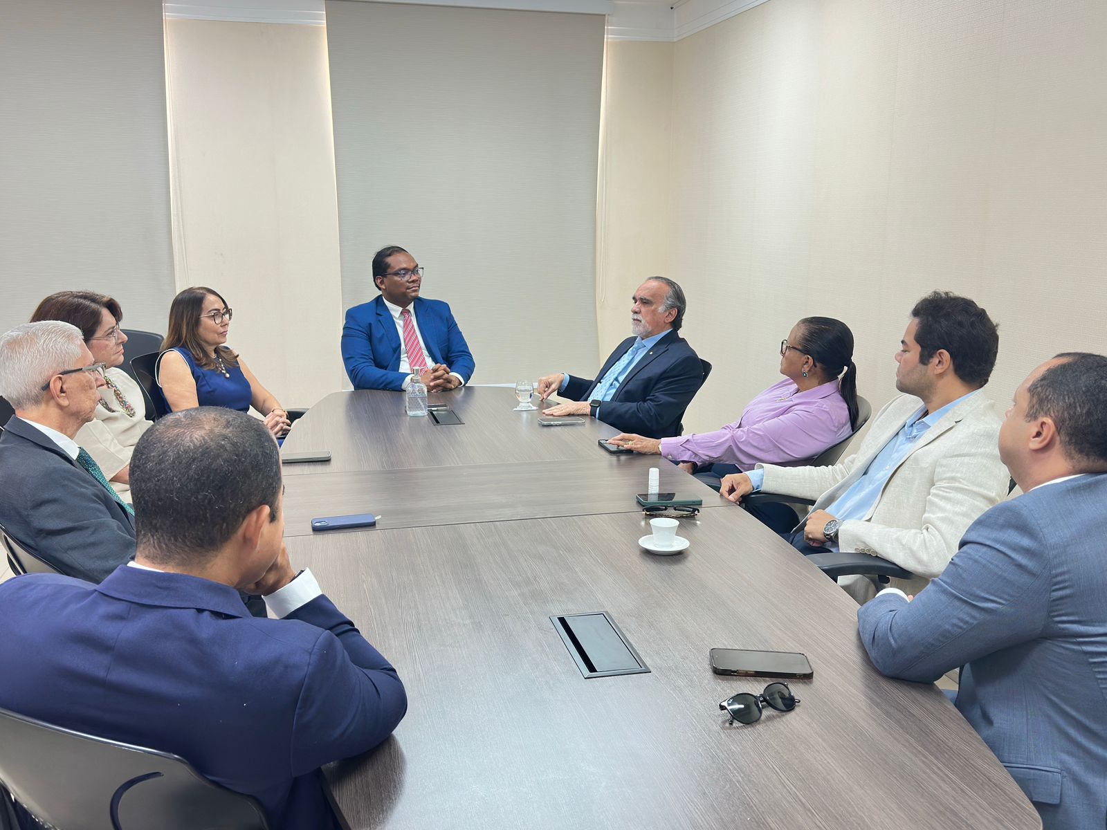 Mostra uma reunião formal em andamento, com as mesmas pessoas sentadas ao redor de uma mesa de reuniões.

O corregedor-geral de Justiça, identificado pelo terno azul e gravata vermelha (o mesmo que aparece no centro na primeira imagem), está à esquerda da mesa, interagindo com os demais presentes.

A conversa parece ser séria e atenta, com todos os olhares voltados para um dos participantes que fala.

Há copos, garrafas de água, xícaras de café e celulares sobre a mesa, indicando um momento de troca institucional.
