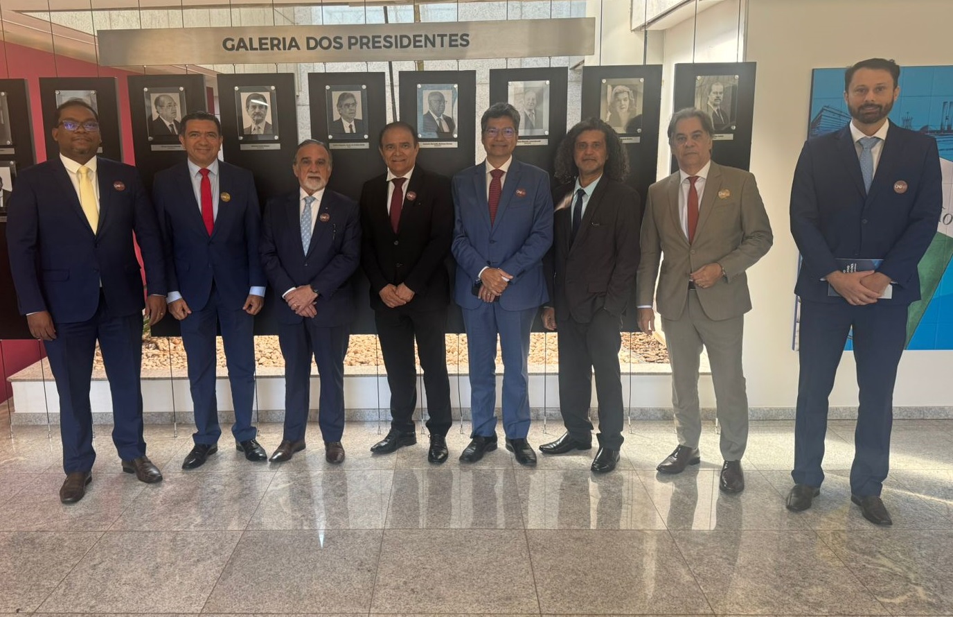 Foto horizontal, colorida, mostra um grupo de homens vestidos de terno, em ambiente interno, posando pra foto diante de um painel com uma galeria de fotos com o nome "Galeria de Presidentes". Todos olham pra frente e alguns têm as mãos cruzadas.