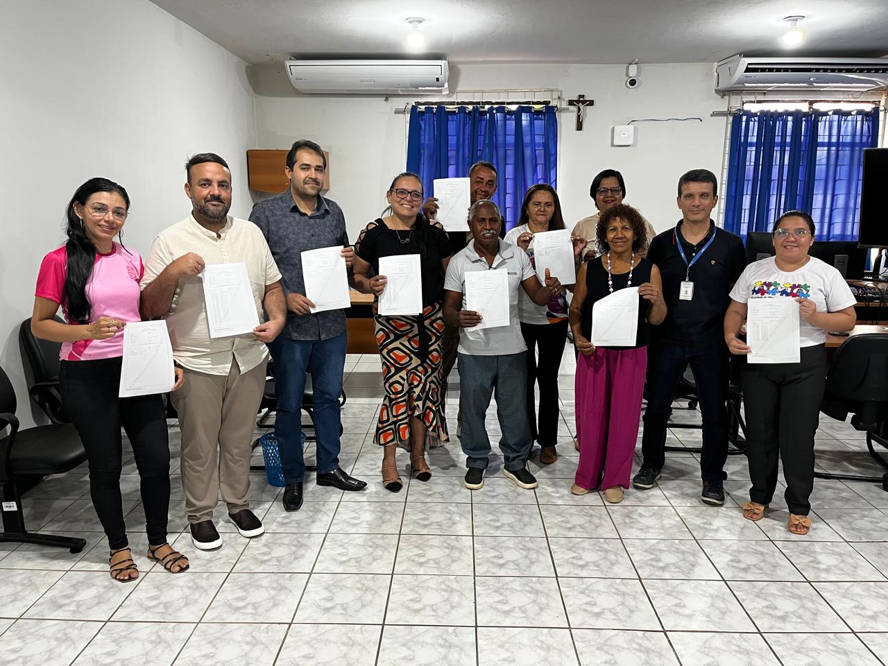 Foto horizontal, colorida, mostra um grupo de 11 pessoas, aparentemente em um escritório ou sala de reunião. Eles estão de pé, dispostos em duas fileiras, e seguram papéis que parecem ser certificados ou documentos. A maioria das pessoas está sorrindo e olhando para a câmera.

As pessoas na foto incluem:

    Uma mulher à esquerda, vestindo uma camiseta rosa e calça preta, segurando um documento.

    Dois homens no centro-esquerda, um com barba e camiseta clara, e outro com camisa escura. Ambos seguram papéis.

    Uma mulher no centro, com óculos e uma blusa estampada, também com um documento.

    Um homem mais velho no centro, com camisa cinza, segurando papéis e com um sorriso.

    Outras pessoas em pé na fila de trás e na extremidade direita, todos segurando documentos.

O fundo da sala tem paredes brancas, um piso de cerâmica branco com rejunte escuro e janelas com cortinas azuis. É possível ver um aparelho de ar condicionado na parede e um crucifixo acima das cortinas. A iluminação parece ser artificial, vinda de lâmpadas de teto.