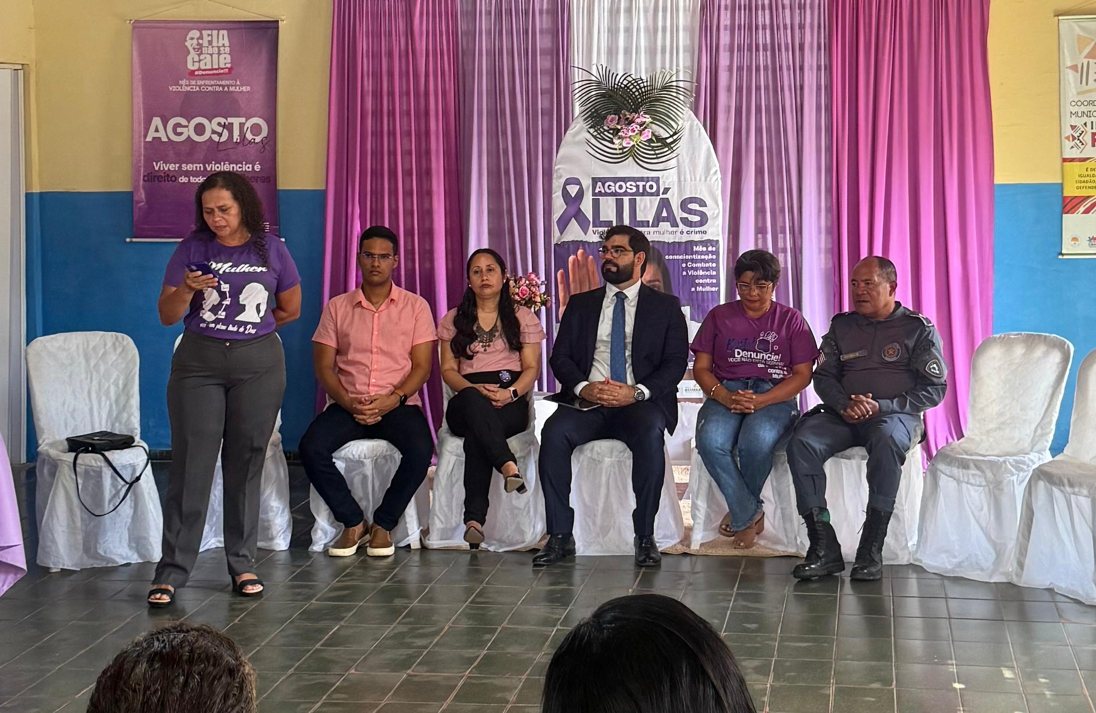 Foto horizontal, colorida, mostra mostra um palestrante em pé no centro, vestindo terno e segurando um tablet, enquanto fala para uma audiência. O palco é simples, com uma cortina roxa e branca ao fundo e um banner roxo com a inscrição "Agosto Lilás", indicando que o evento está relacionado a alguma campanha de conscientização sobre violência contra a mulher. A audiência, composta majoritariamente por mulheres, está sentada em cadeiras brancas de plástico. Na frente, há um pequeno grupo de pessoas também sentadas, mas em um semicírculo.