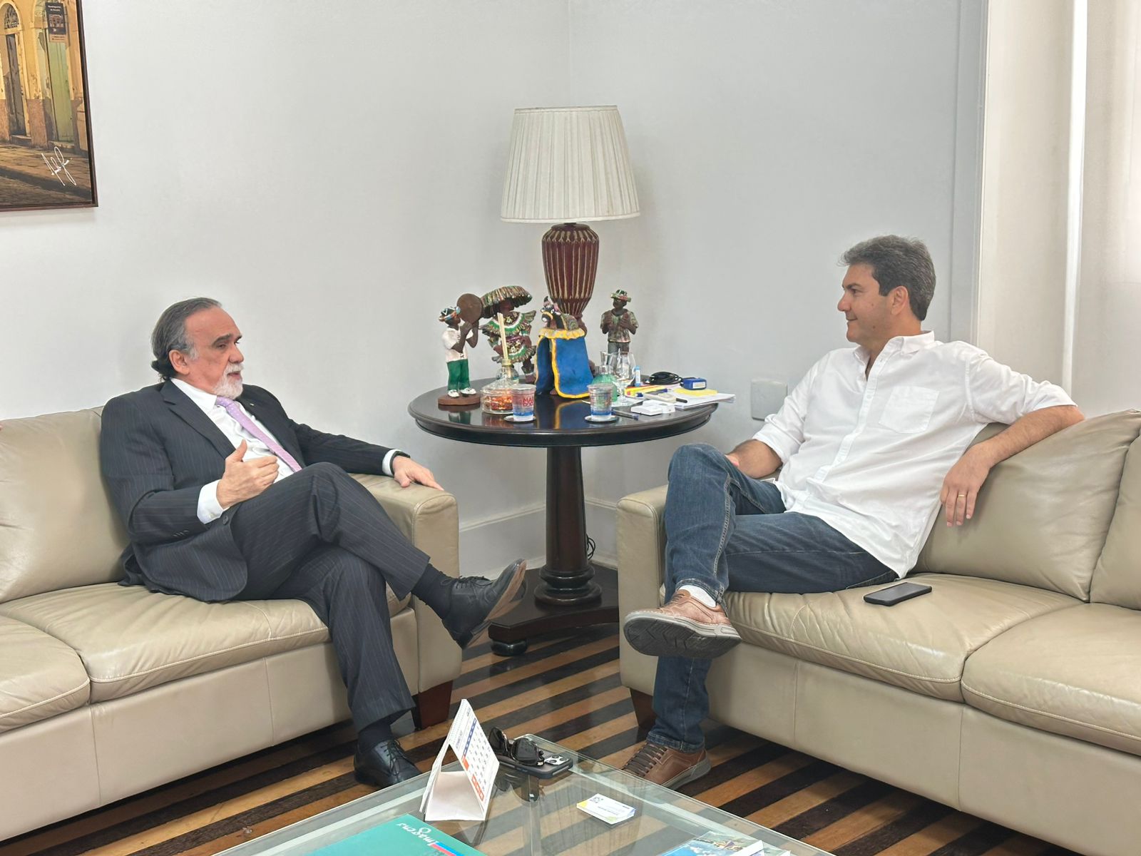 Dois homens estão sentados em sofás opostos, conversando.

À esquerda, o homem de terno escuro (o mesmo da primeira foto) fala gesticulando com a mão.

À direita, o homem de camisa branca e jeans (também da primeira foto) ouve atentamente, com o braço apoiado no encosto do sofá.
Entre eles, há uma mesa lateral com esculturas decorativas, uma luminária e alguns objetos pessoais. O cenário transmite um encontro institucional em clima descontraído.