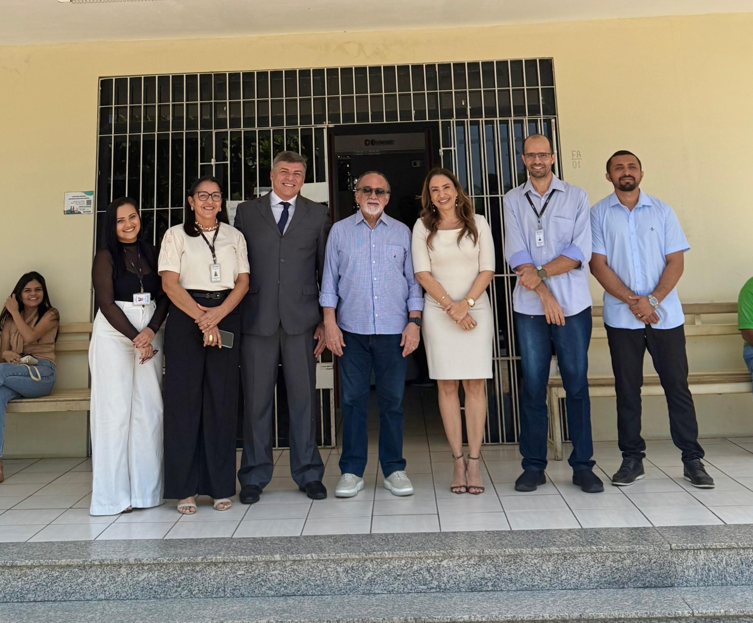 Grupo de sete pessoas em pé, posando para foto em frente a um prédio. Estão lado a lado, sorrindo, alguns de crachá.