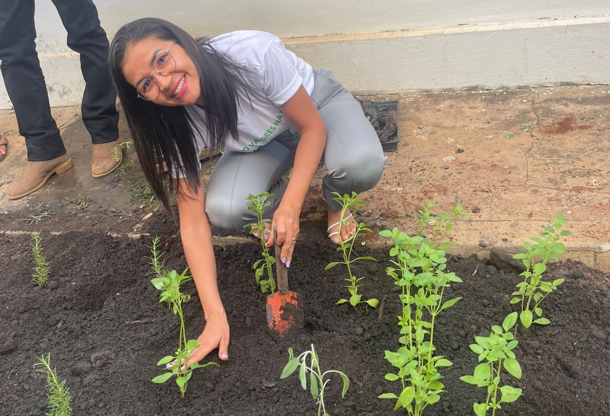 Foto horizontal, colorida, mostra uma mulher sorridente, usando óculos e uma camiseta branca, está agachada plantando mudas de ervas em um canteiro de terra preta, usando uma pequena pá. Há detalhe de pernas outra pessoa parcialmente visível à esquerda, de calça jeans azul.