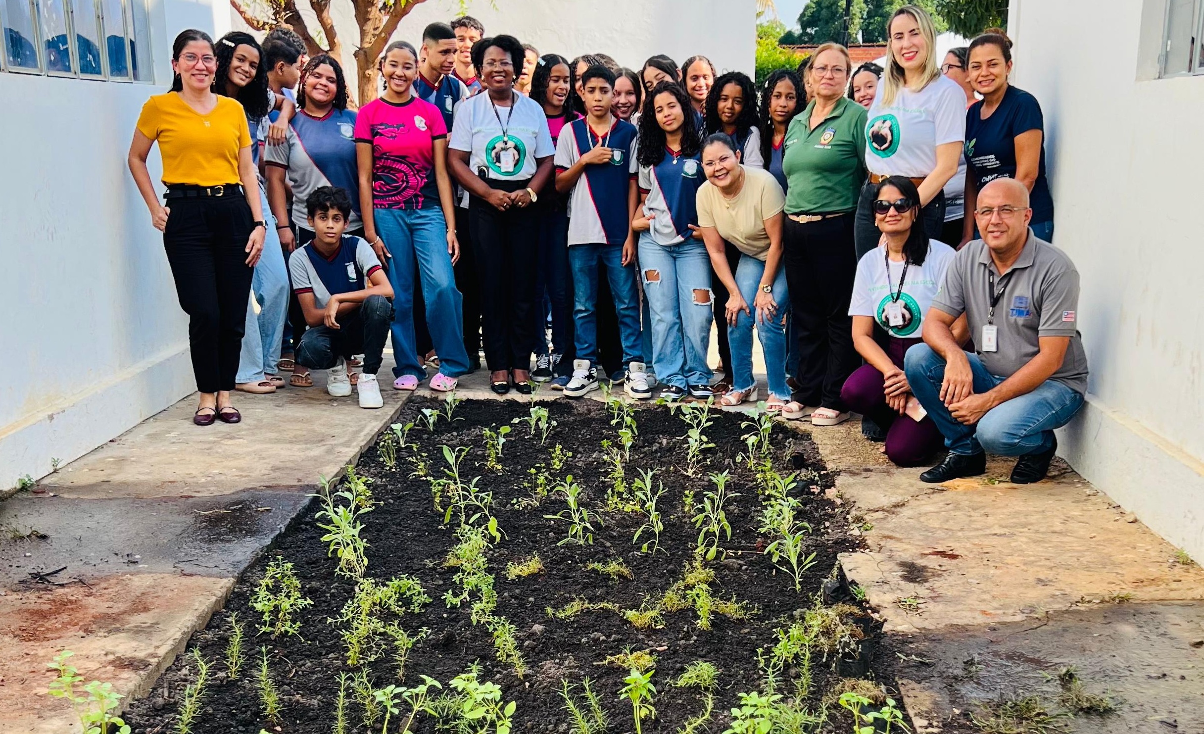 Foto horizontal, colorida, de um grupo grande de pessoas, incluindo jovens e adultos, posando para uma foto em um pátio claro, com paredes brancos na lateral, céu azul. Em primeiro plano, há um canteiro retangular recém-plantado com várias mudas verdes.