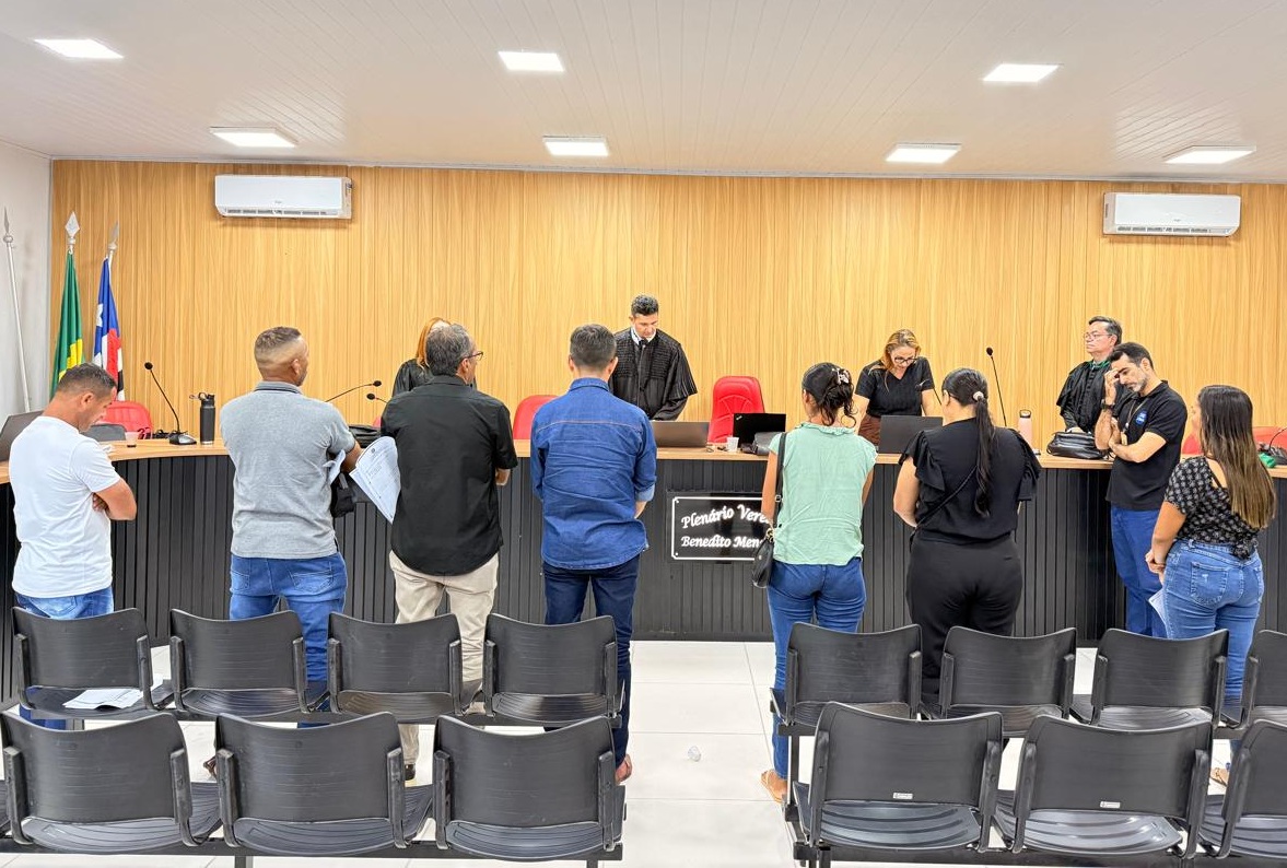 Foto horizontal, colorida, mostra oito pessoas (cinco homens e três mulheres) estão de pé no primeiro plano, de costas ou parcialmente de perfil para a câmera, voltadas para o que parece ser o corpo de jurados e o juiz. 

Na parte de trás, elevada em um estrado escuro, está uma longa bancada preta. Atrás dela, estão sentados ou em pé pelo menos quatro ou cinco indivíduos, que parecem ser os oficiais do tribunal:

Ao centro: Um homem de toga preta está em pé, no centro da bancada, provavelmente o Juiz Presidente do julgamento.

Nas laterais, outras pessoas estão sentadas ou em pé, também vestidas formalmente (algumas de preto), possivelmente advogados, promotores, ou assistentes.

Na parte inferior da imagem, há várias fileiras de cadeiras plásticas escuras vazias, destinadas ao público ou aos jurados (se estiverem em pé no momento).