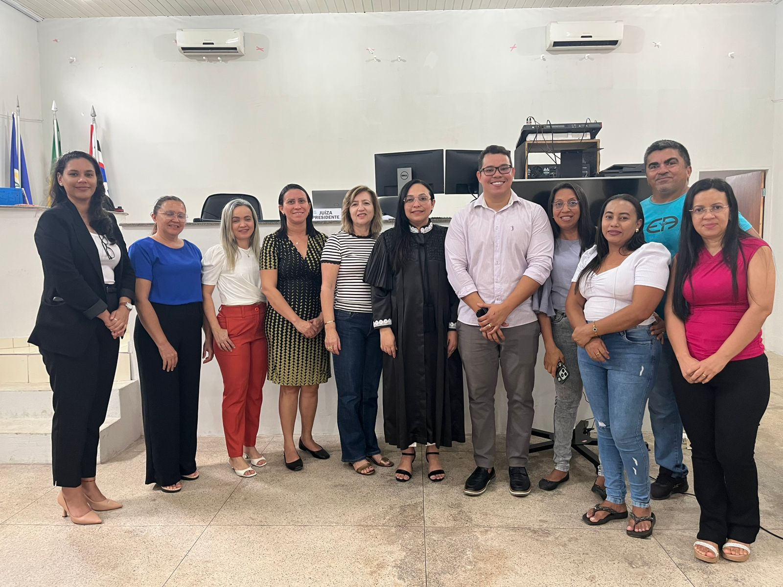 Foto horizontal, colorida, mostra um grupo de 11 pessoas posando para uma foto em um ambiente que parece ser um tribunal ou fórum jurídico no Brasil. O grupo é composto por dez mulheres e um homem, todos em trajes profissionais ou casuais. Uma das mulheres, ao centro, está vestindo uma toga preta, indicando ser uma juíza ou magistrada.
Ambiente: O local possui características de uma sala de audiência ou gabinete de tribunal, incluindo:
Um balcão ou mesa de trabalho ao fundo.
Cadeiras de escritório e uma placa com a inscrição "SALA AUDIÊNCIA".
Bandeiras (uma do Brasil e outra que pode ser de um estado ou município) visíveis no lado esquerdo.
Equipamentos eletrônicos, como caixas de som e aparelhos de ar-condicionado na parede superior.