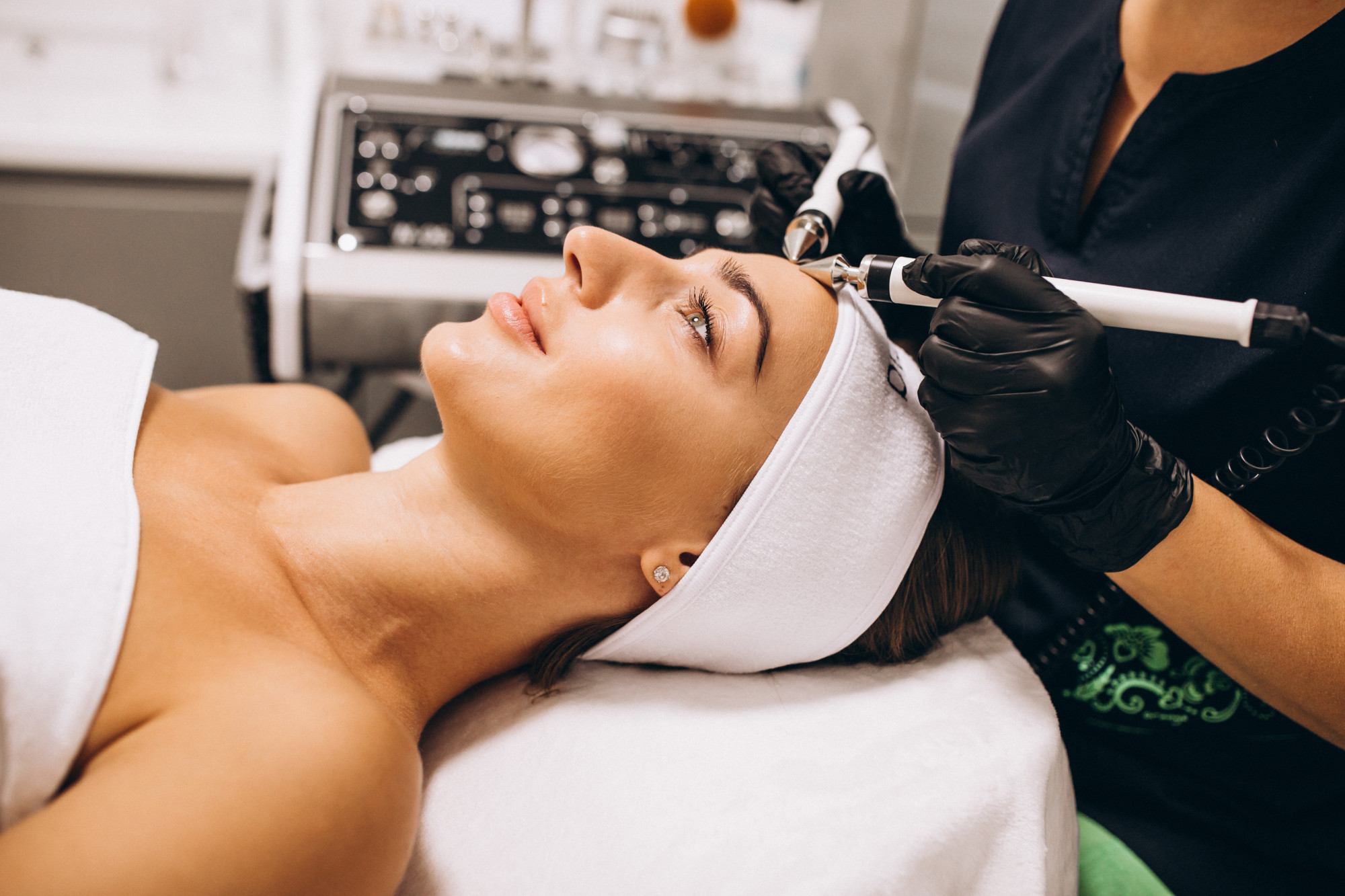 Foto horizontal, colorida, mostra uma mulher branca, cabelos pretos rpesos com faixa branca, deitada enquanto recebe um tratamento facial. à direita, detalhe de pessoa branca, de jaleco preto, que realiza o procedimento está usando luvas pretas e segurando um dispositivo branco próximo ao rosto da mulher deitada. Ao fundo, há uma máquina com diversos botões e controles.