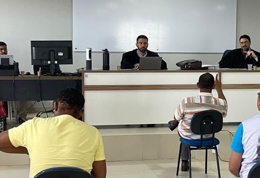 imagem na qual aparece um juiz, ao longe, sentado, e outras pessoas de costas, em uma sala, durante uma sessão de julgamento