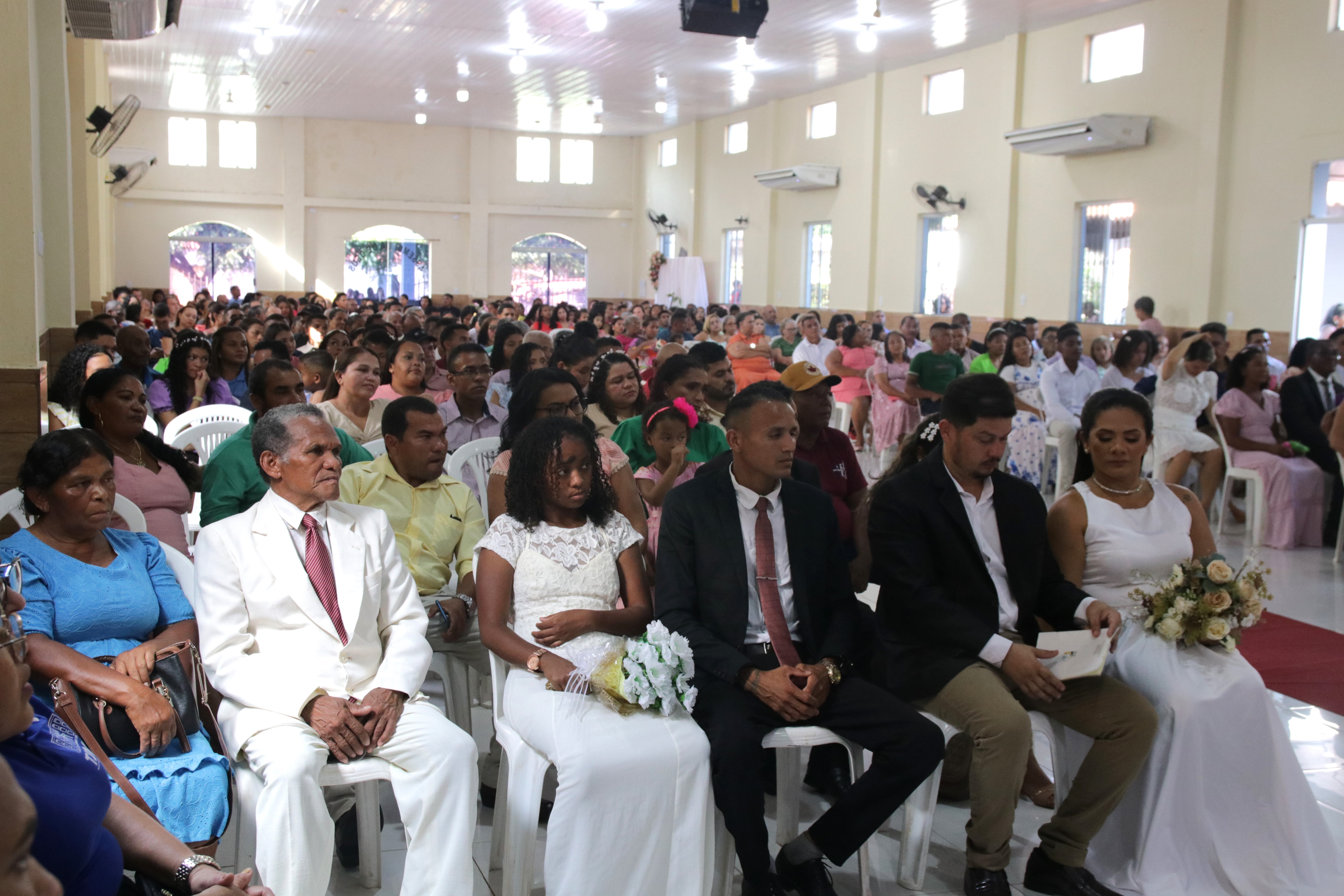 Foto horizontal, colorida, de homens e mulheres sentados em duas fileiras de cadeiras em salão de Igreja, divididas por um tapete vermelho. Algumas mulheres estão vestidas de vestidos brancos e seguram buquê de flores. Ao fundo, paredes na cor amarela-clara, com janelas e luminárias.