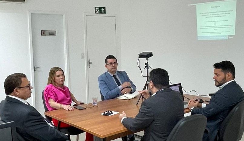 Foto horizontal, colorida, de quatro homens e uma mulher reunidos em torno de mesa de madeira, em sala de paredes e chão claros, em ambiente interno. Sobre a mesa há equipamentos como computador, projetor e tripé. Ao fundo, projeção de tele de computador na parede.
