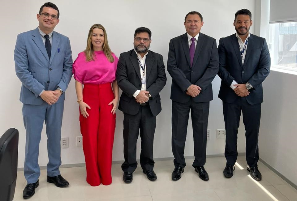 Foto horizontal, colorida, de homens e uma mulher de pé, lado a lado, diante de parede clara, posando para foto, em roupas formais e ambiente interno. Do lado direito, persiana cobrindo metade de janela.