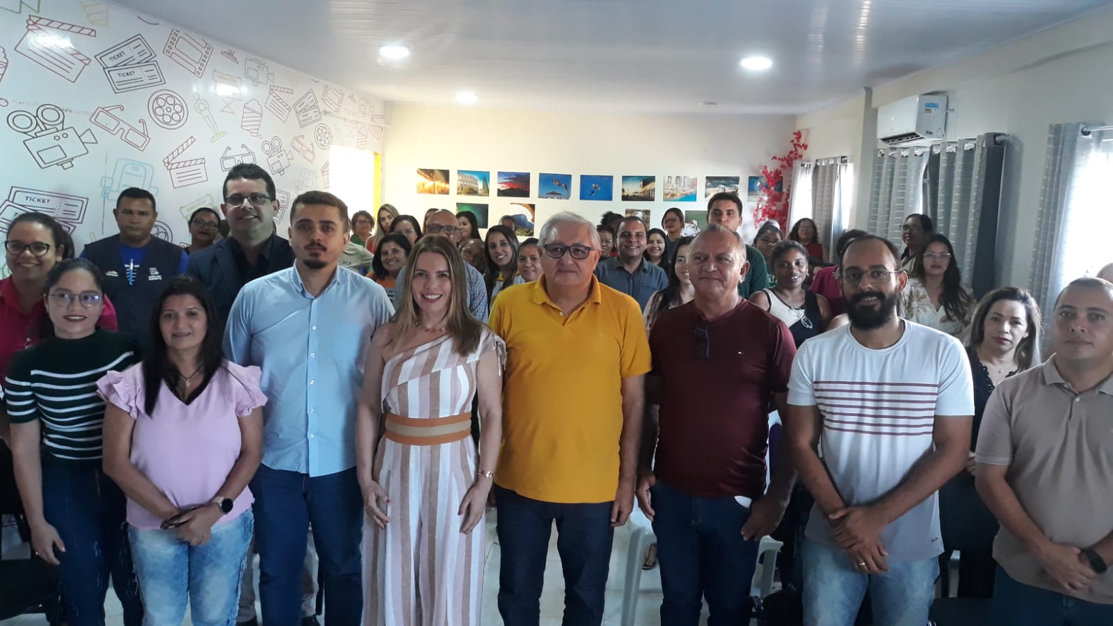 Foto horizontal, colorida, de homens e mulheres de pé, em auditório de paredes e chão claros, vestidos com roupas formais.À esquerda, parede coberta com adesivo de fundo branco com desenhos de equipamentos de cinema. À direita, três janelas cobertas com cortinas claras listradas. Ao fundo, parede branca com quadros coloridos.